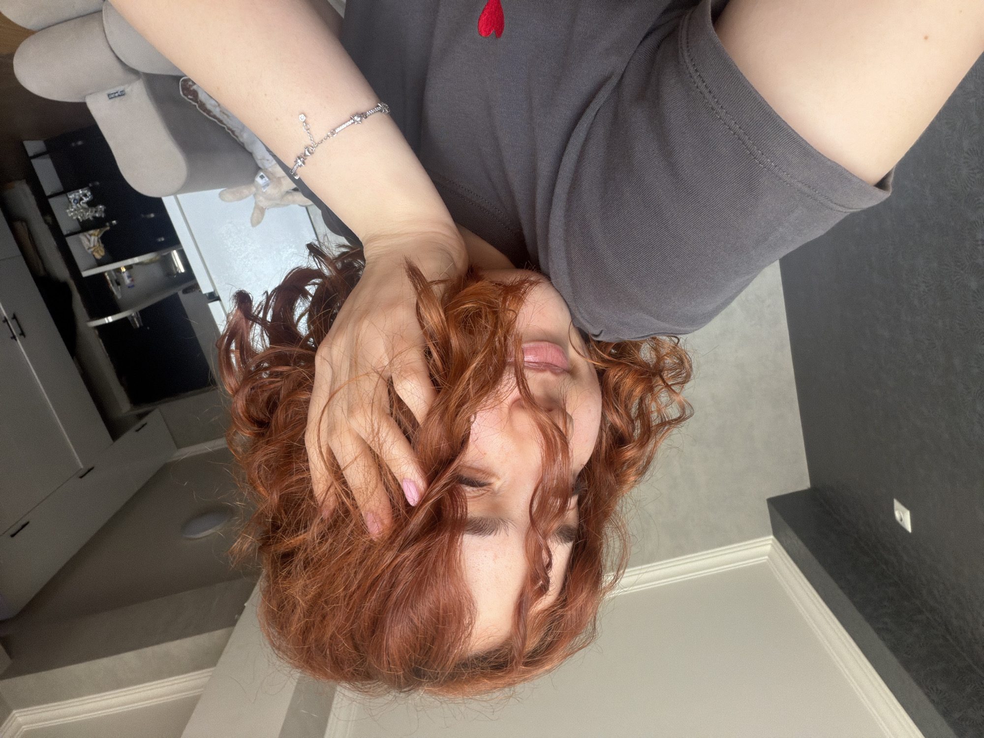 A fair-skinned individual with red curly hair, wearing a dark gray shirt adorned with a red heart, is partially visible in a vertically inverted orientation. Their face is partially obscured by their hand, which has pink nail polish. A silver bracelet is visible on their wrist. The backdrop includes a gray sofa, black and white cabinets, a white wall with molding, and a dark gray surface. It is not possible to determine if the location is in Tbilisi, Georgia.