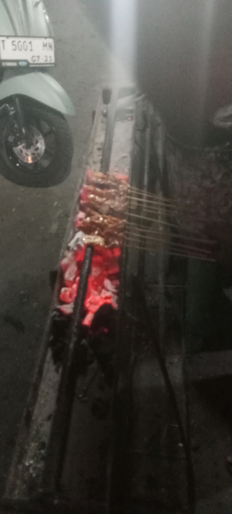 The image shows a close-up of several skewers of meat being grilled over hot coals. The meat appears to be dark brown and is arranged in rows on metal skewers. The coals are glowing red and are situated in a metal grill. In the background, to the left, is a portion of a light-colored motorcycle, including its front wheel and a license plate that reads "T 5001 MN 07-21". The overall scene suggests an outdoor food stall or a casual outdoor cooking setting, possibly in Karawang, Indonesia, given the license plate format and the presence of street food preparation. The lighting is dim, and the image is slightly blurred, indicating it was taken in low light conditions, possibly at night.