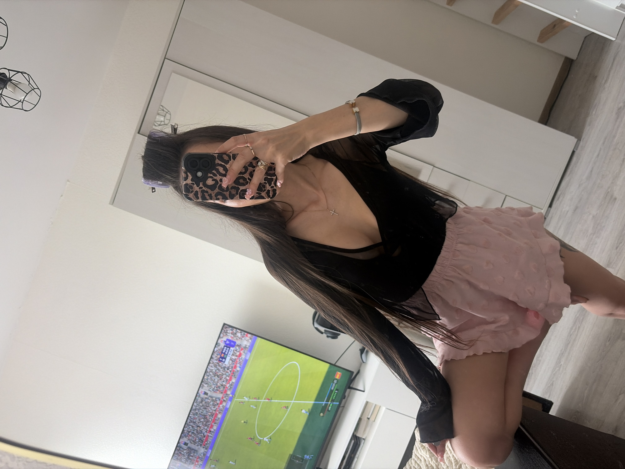 A person is depicted taking a mirror selfie in a domestic interior. The subject, appearing female, has long dark hair styled with two purple hair rollers on top. She is wearing a sheer black long-sleeved top over a black bra and pink, textured, ruffled shorts. Her right hand, adorned with multiple rings and manicured nails, holds a leopard-print smartphone that covers her face. A gold cross necklace is visible on her chest, and a light-colored bracelet is on her left wrist. She is in a crouching position on a dark upholstered surface, partially covered by a cream-colored textured throw.
The background, visible through the mirror's reflection, includes a white wall and a large flat-screen television on a white low-profile media console. The TV displays a soccer video game with a 0:0 score and team names in a non-Latin, Cyrillic-like script. A gaming headset is also visible on the console. To the right, a white multi-panel wardrobe or closet is present. Overhead, a black wireframe ceiling light fixture with two exposed bulbs is visible. The flooring is a light-colored wood or laminate. A secondary, tall, vertical mirror with light wooden accents is partially visible on the far right. No discernible features definitively indicate the location as Saint Petersburg, Russia.