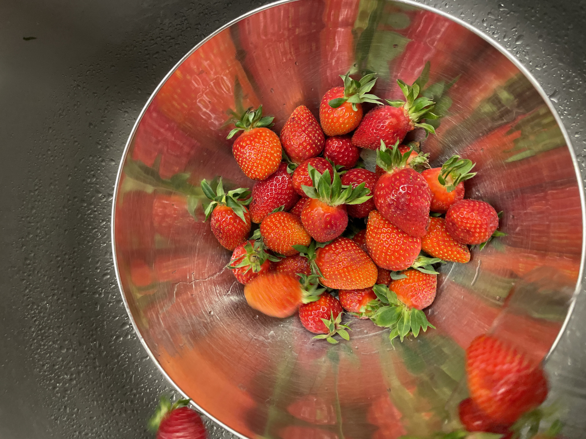 A stainless steel bowl filled with ripe strawberries, some with their green leafy stems still attached, sits on a dark, wet surface. The interior of the bowl is reflective, appearing distorted due to the curved surface. The dark surface surrounding the bowl has a wet or moist appearance. The image suggests a clean, possibly kitchen environment where fruit has just been washed. The city of Boreti, Montenegro is not discernible within the image.