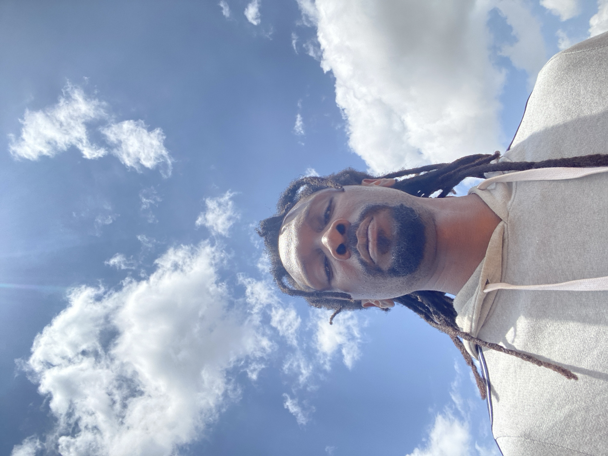 A low-angle selfie, looking up at a Black man against a bright blue sky with scattered white clouds. The man has dark dreadlocks and a beard, and is wearing a light-colored hooded sweatshirt. The image appears to be taken outdoors, with the sun high in the sky, causing some glare. There is no indication of the specific location in Gura Top, Nigeria.