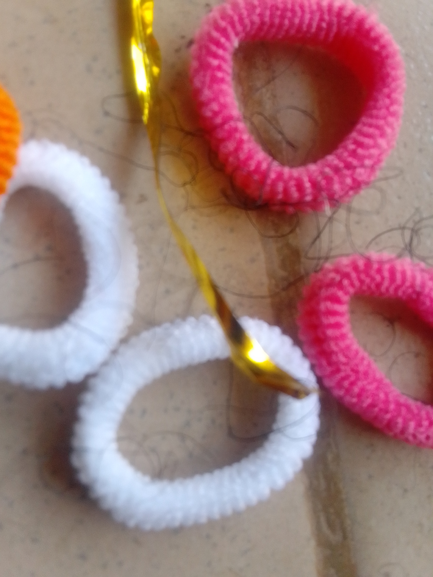 A macro shot shows several fabric scrunchies and a gold ribbon on a neutral surface. Two pink scrunchies are visible, one larger and to the top right of the frame, and a smaller one to the bottom right. Two white scrunchies are present, one to the left of the frame and another below it, overlapping slightly. A thin, gold, shiny ribbon runs diagonally across the center of the image. The surface beneath appears to be a textured, beige or light brown material, possibly tile or linoleum, with small dark specks. The lighting is somewhat dim, creating soft shadows. There are also several dark, thin strands scattered across the scrunchies and surface, which appear to be hair.