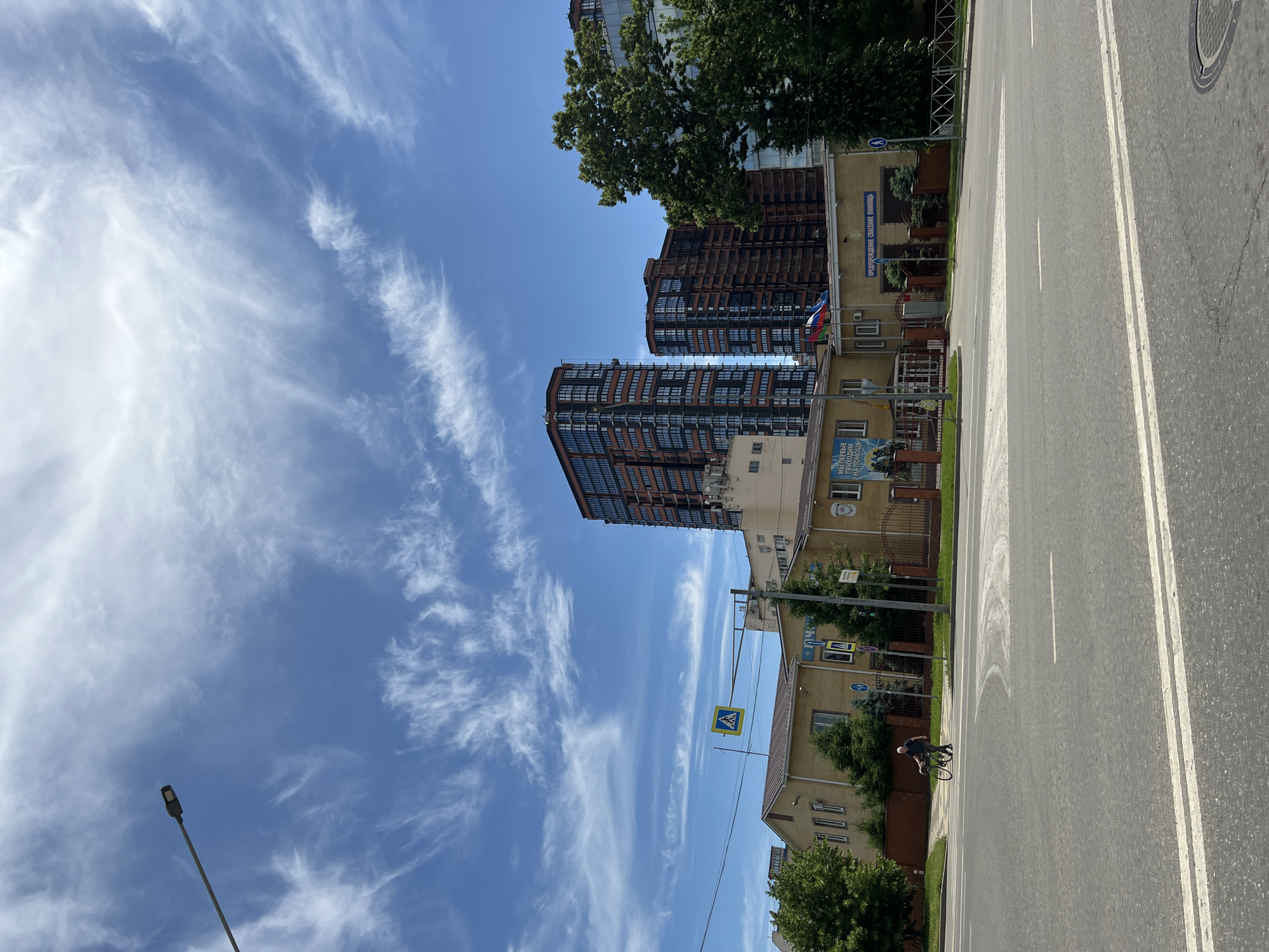 The image captures an urban street scene in Krasnodar, Russia, under a bright, clear blue sky adorned with wispy, white cirrus clouds, indicating a sunny midday or early afternoon.
In the foreground, a wide asphalt road stretches into the distance, marked with crisp white lane lines, including a double solid line separating traffic directions. Along the right side of the road, a person wearing a dark t-shirt and a light-colored cap or helmet is riding a bicycle, moving away from the viewer.
On the left side of the road, several buildings are visible. In the background, two tall, modern-looking buildings with dark, grid-patterned window facades rise prominently. Closer to the foreground is a lower, yellowish building with a fence in front. This building features multiple windows and displays Cyrillic text on its facade: "ПРЕДУПРЕЖДЕНИЕ СПАСЕНИЕ ПОМОЩЬ" (WARNING RESCUE HELP) and "МЫ ПЕРВЫЕ ПРИХОДИМ НА ПОМОЩЬ" (WE ARE THE FIRST TO COME TO HELP), suggesting it is a public safety or emergency services facility. Three small flags, including the Russian flag, are mounted on this building.
Green trees with dense foliage are interspersed throughout the scene, providing shade and natural elements around the buildings. A pedestrian crossing sign (blue square with a white triangle and pedestrian symbol) is suspended over the road. On the far left, a tall, dark street light pole stands out against the bright sky. The overall scene conveys a typical, active day in an urban environment.