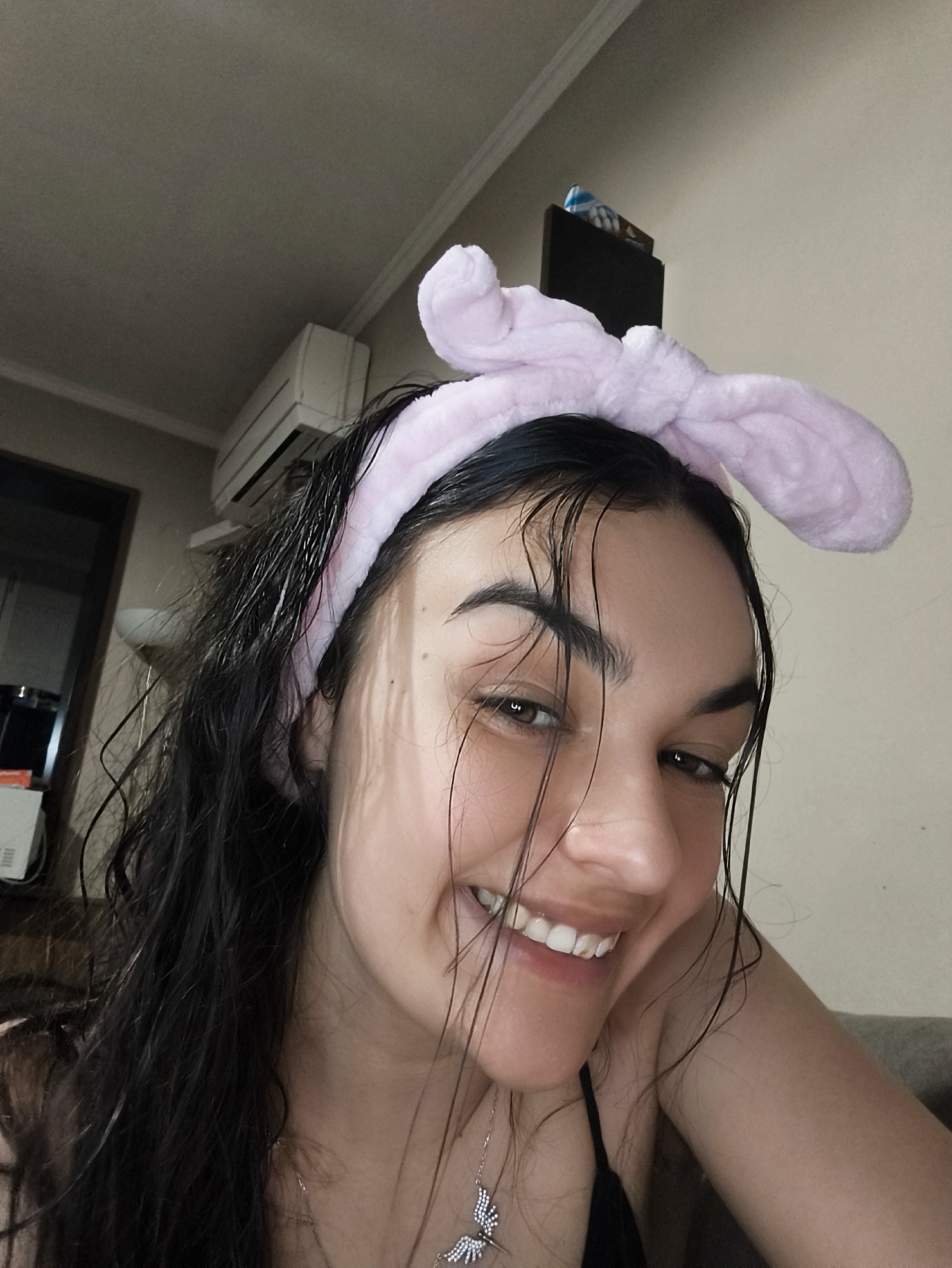 A young woman with wet, dark hair, wearing a pink fuzzy headband with rabbit ears, smiles broadly at the camera. She is wearing a black top and a delicate silver necklace with a butterfly pendant. The background shows an indoor setting with an air conditioning unit on the wall and a dark rectangle on the wall, possibly a TV. The lighting suggests it is daytime. The image appears to have been taken inside an apartment in Sofia, Bulgaria.