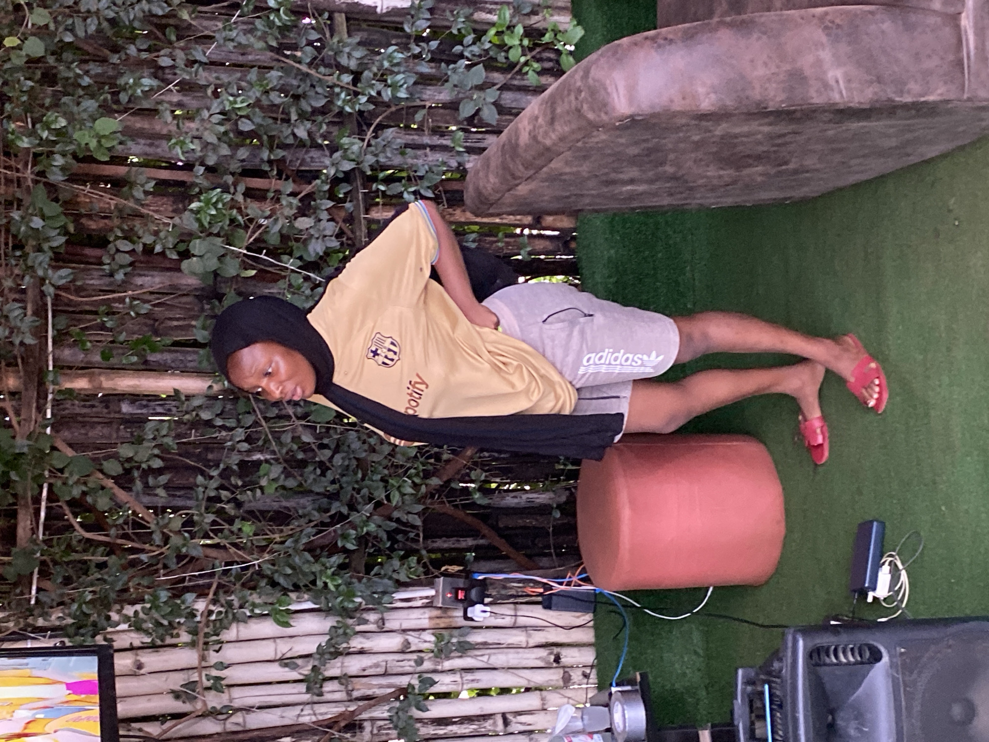 A young Black woman wearing a black hijab, a gold t-shirt with a Barcelona logo and "spotify" printed on it, and grey Adidas athletic shorts stands with her hand on her hip. She is facing to her left, looking up and to the left. She is wearing red slip-on sandals. The woman is standing on a green artificial turf floor in an outdoor area. Behind her is a wooden bamboo fence covered in green foliage. To her left, there is a red cylindrical ottoman, a black speaker, and a power strip with cables. To her right, a portion of a brown, worn leather sofa is visible. The image appears to have been taken in the city of Gura Top, Nigeria.