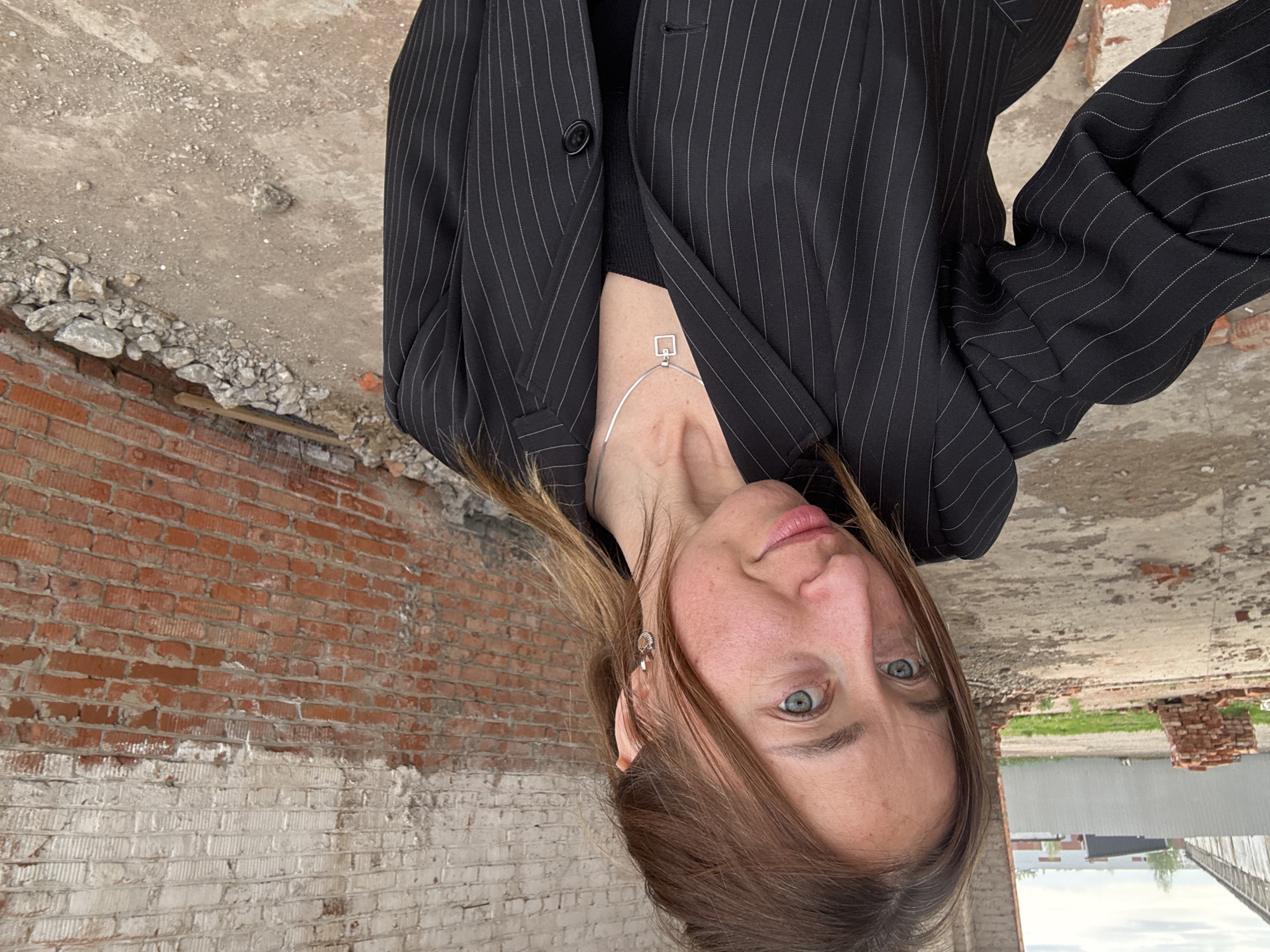 A woman with light brown hair and blue eyes is seen in an upside-down perspective. She is wearing a black pinstripe blazer over a black top and a silver necklace. The location is an abandoned structure in Chaikovsky, Russia, based on prior data, featuring brick walls and dilapidated concrete surfaces. The viewpoint is from the floor looking upward at the woman.