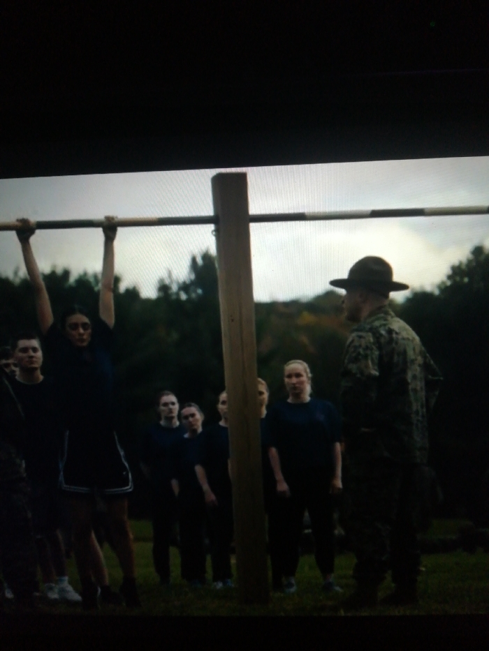 A group of people in dark clothing are being trained. One person hangs from a pull-up bar. Several others stand in line behind her. A man in military camouflage uniform and a wide-brimmed hat faces the group. The background includes trees and a grassy area. The light in the image is dim.