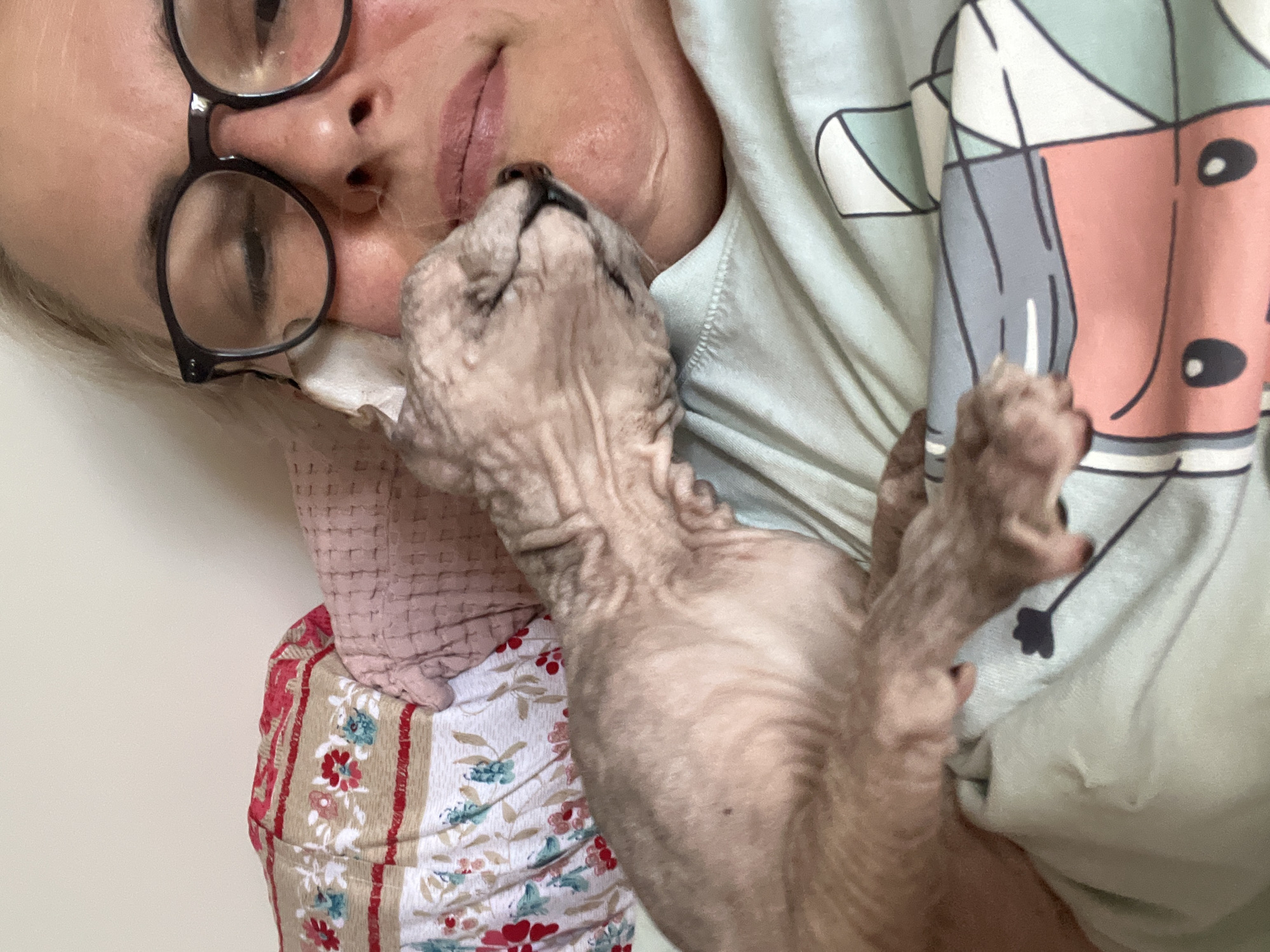 The image shows a close-up of a woman with blonde hair and glasses lying down, cuddling a hairless cat. The cat is nudging the woman's face. The background consists of bedding with floral patterns and a light-colored wall. The scene appears to be indoors, possibly in a bedroom. There is no information to indicate the location in the city of Korolyov, Russia.