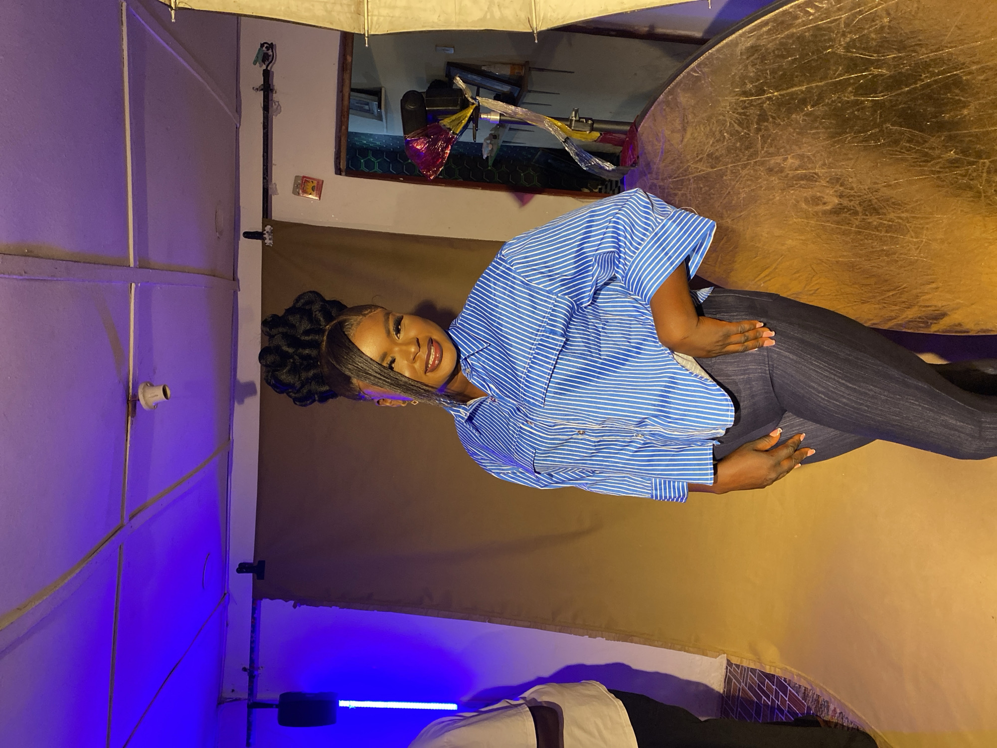 The image shows a woman with dark skin and a high braided hairstyle wearing a blue and white striped shirt and dark jeans. She is smiling at the camera. She is positioned in front of a backdrop that transitions from brown to purple. A large gold reflector is positioned behind her on the right. The scene appears to be in a studio setting with lighting equipment visible, including an umbrella light modifier. A partial view of someone's upper body is seen in the lower portion of the frame. The location in Vom, Nigeria is not definitively established in the image.