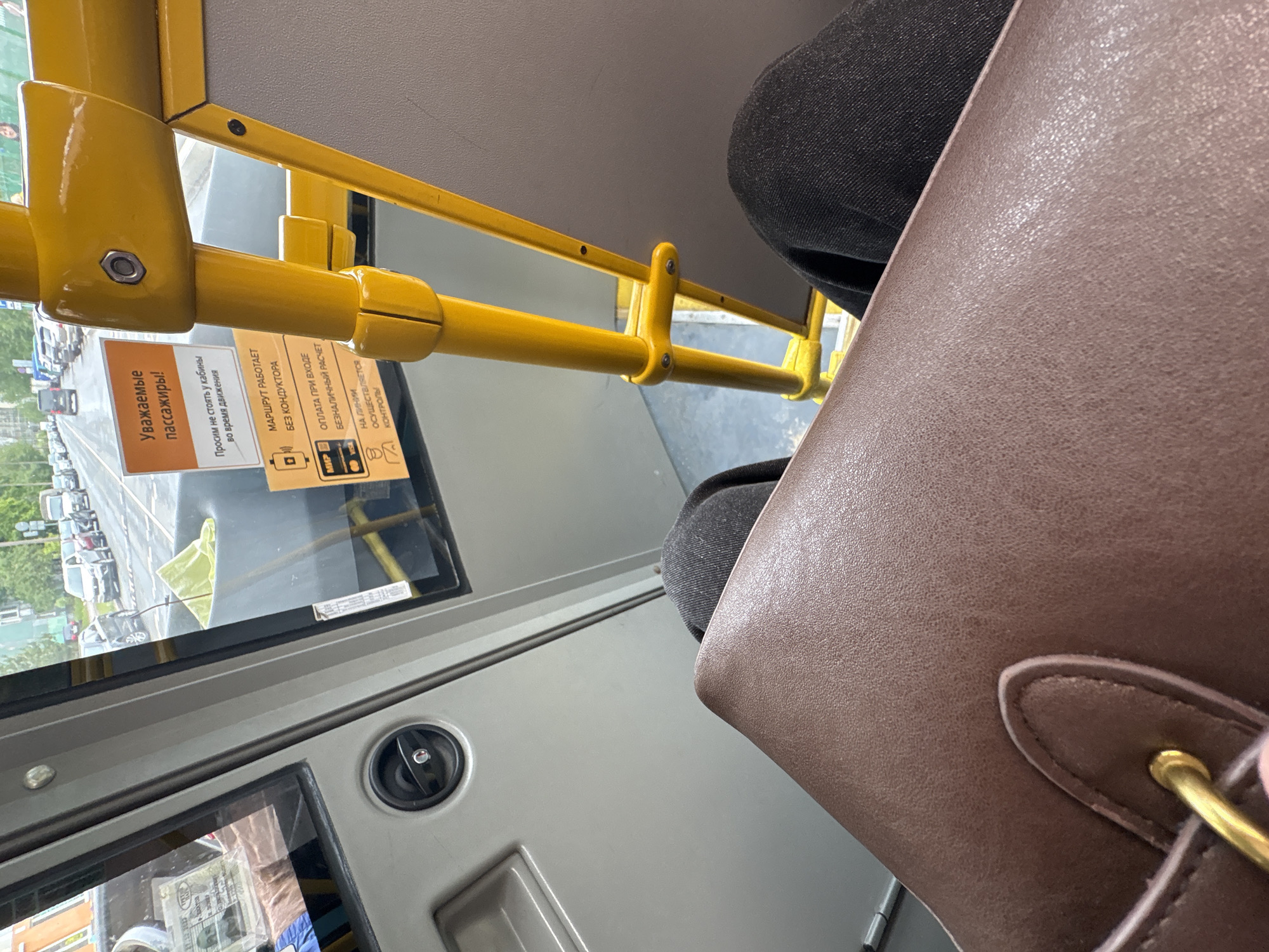 The image, likely taken in Saint Petersburg, Russia, shows an interior view of a bus. A brown leather bag is prominently positioned in the foreground. The bus features yellow handrails and grey paneling. Through the bus window, a cityscape with parked cars and greenery can be seen. Warning signs in Russian are affixed to the bus window, and a payment console is visible. A passenger wearing black denim jeans is also in the frame.