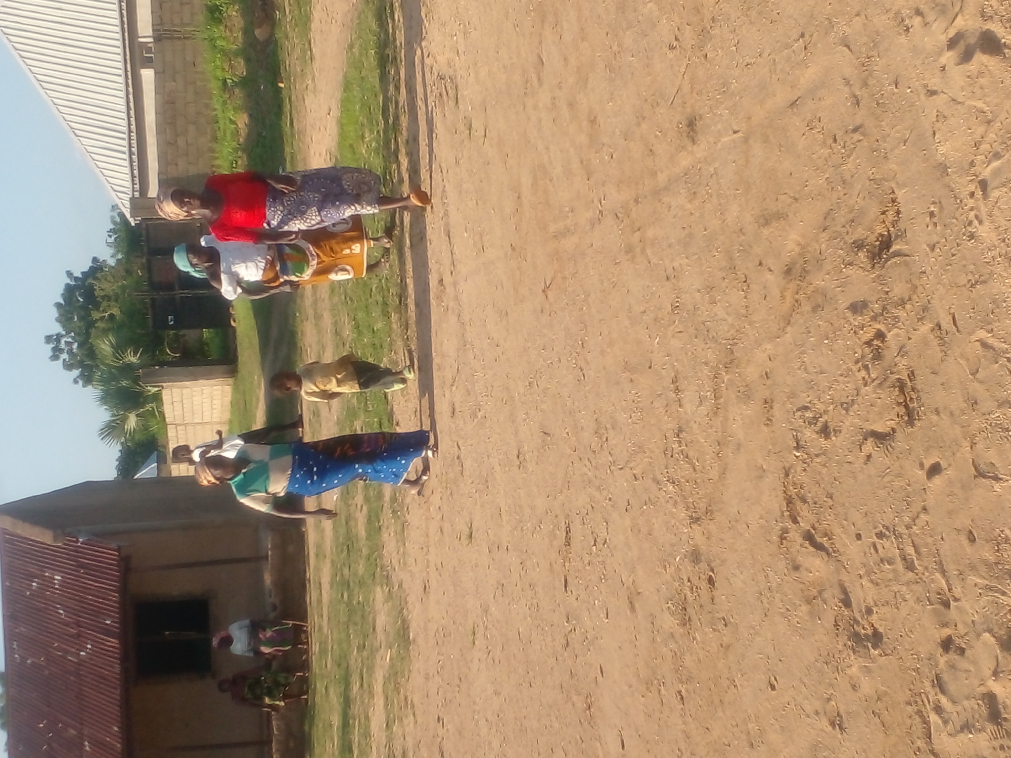 In Langtang, Nigeria, four individuals are present in an outdoor, unpaved area. A child, wearing a light-colored shirt and dark pants, stands in the foreground. To the left, an adult woman, identified as wearing a blue and white striped shirt with a blue and white patterned skirt, is walking. Two other adult women are also visible. One, in the mid-ground, wears a white top and a patterned skirt with a yellow and brown design. The other, in the background, is dressed in a red top and a patterned skirt in shades of blue and purple. The scene is characterized by a sandy ground with some sparse grass. In the background, a simple, unpainted building with a corrugated metal roof is situated. Further back, another building with a lighter, possibly metal, roof and a brick wall are visible, along with some greenery and trees. The overall environment suggests a rural or semi-urban setting.