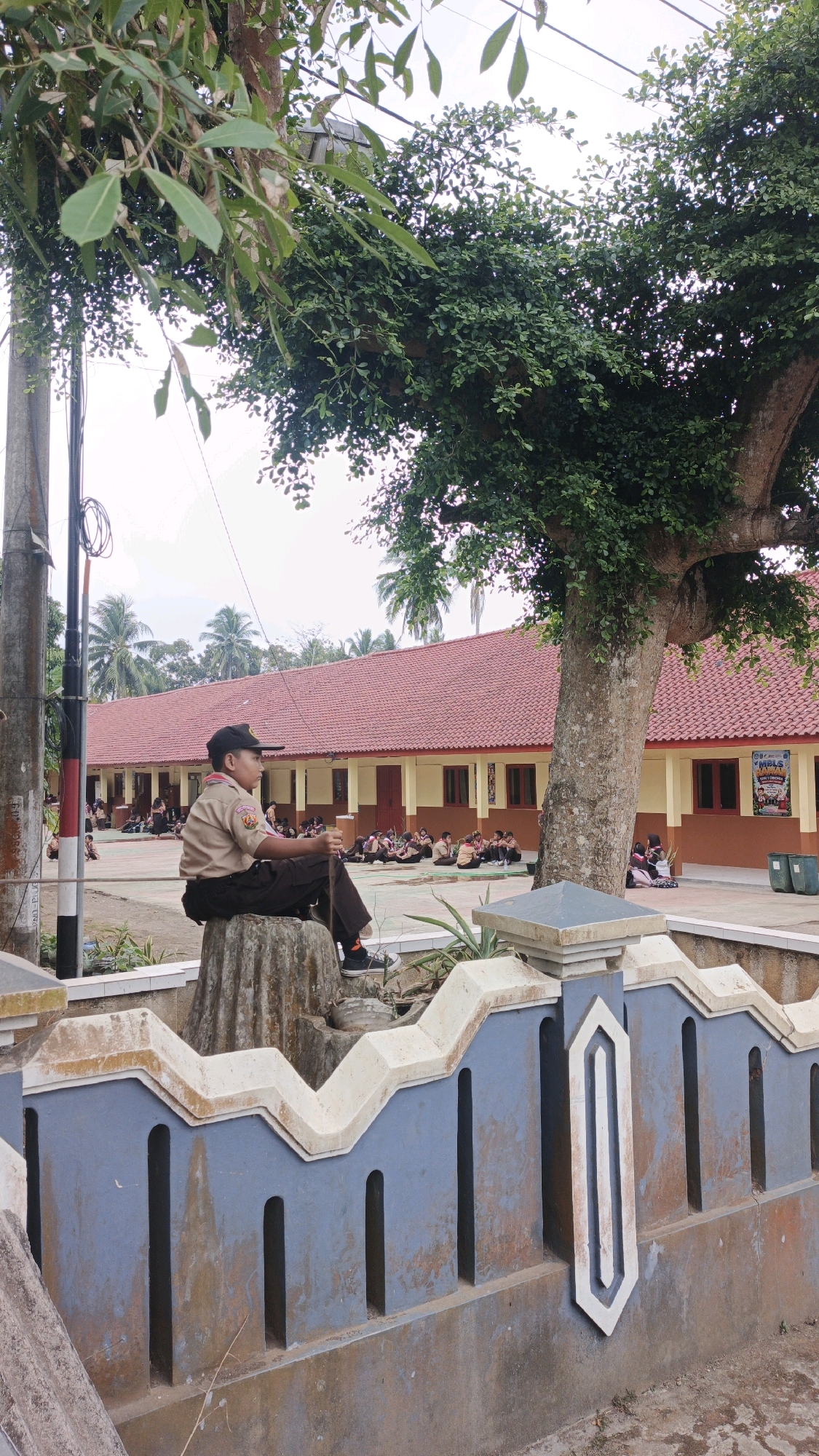 A young Scout, wearing a tan uniform and a dark cap, sits on a carved stone structure in front of a school building in Cibenda, Indonesia. The Scout is holding a dark stick. Behind him, several other Scouts are seated on the ground in a courtyard. The school building has a red tiled roof and pale yellow walls with brown trim. A large tree with dense green foliage looms over the scene to the right. A blue and white decorative fence separates the foreground from the courtyard. The sky is overcast.