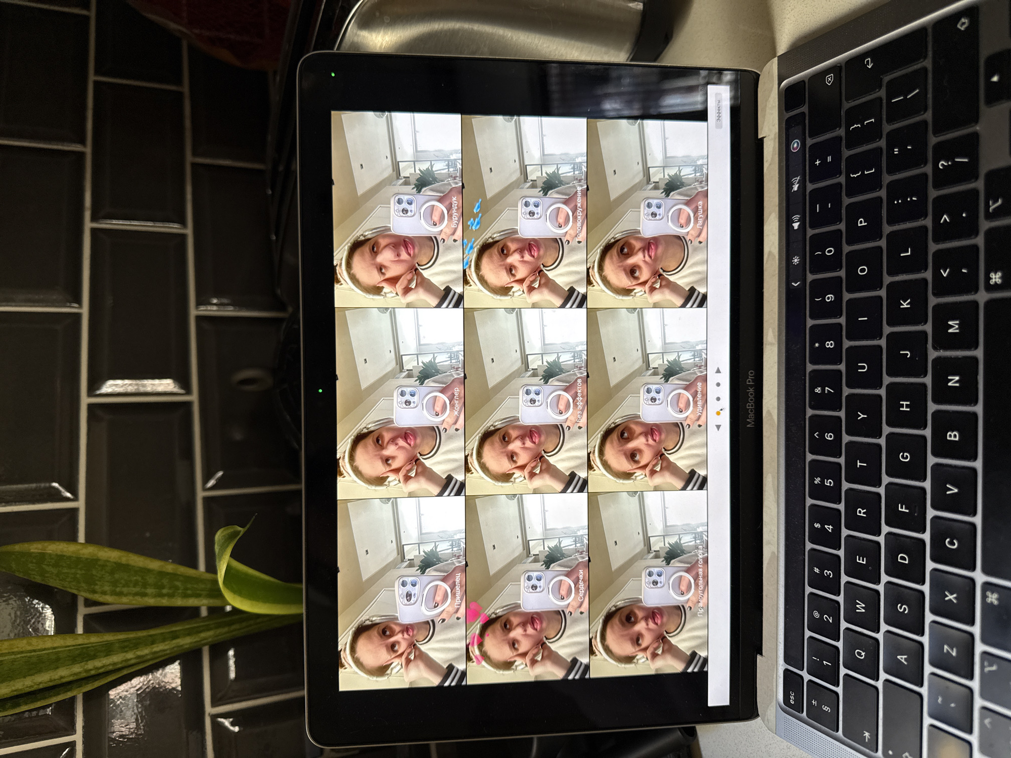 A high-angle, indoor shot depicts a portion of a laptop displaying a grid of nine video previews of a young woman holding a phone with a ring light. Each video shows the woman posing with a different filter effect displayed on the phone screen. The laptop is sitting on a black tile countertop beside a snake plant. The edge of the stainless steel sink is visible in the background. The laptop is a MacBook Pro and the keyboard is visible. The city of Barcelona, Spain is not detectable from this image.