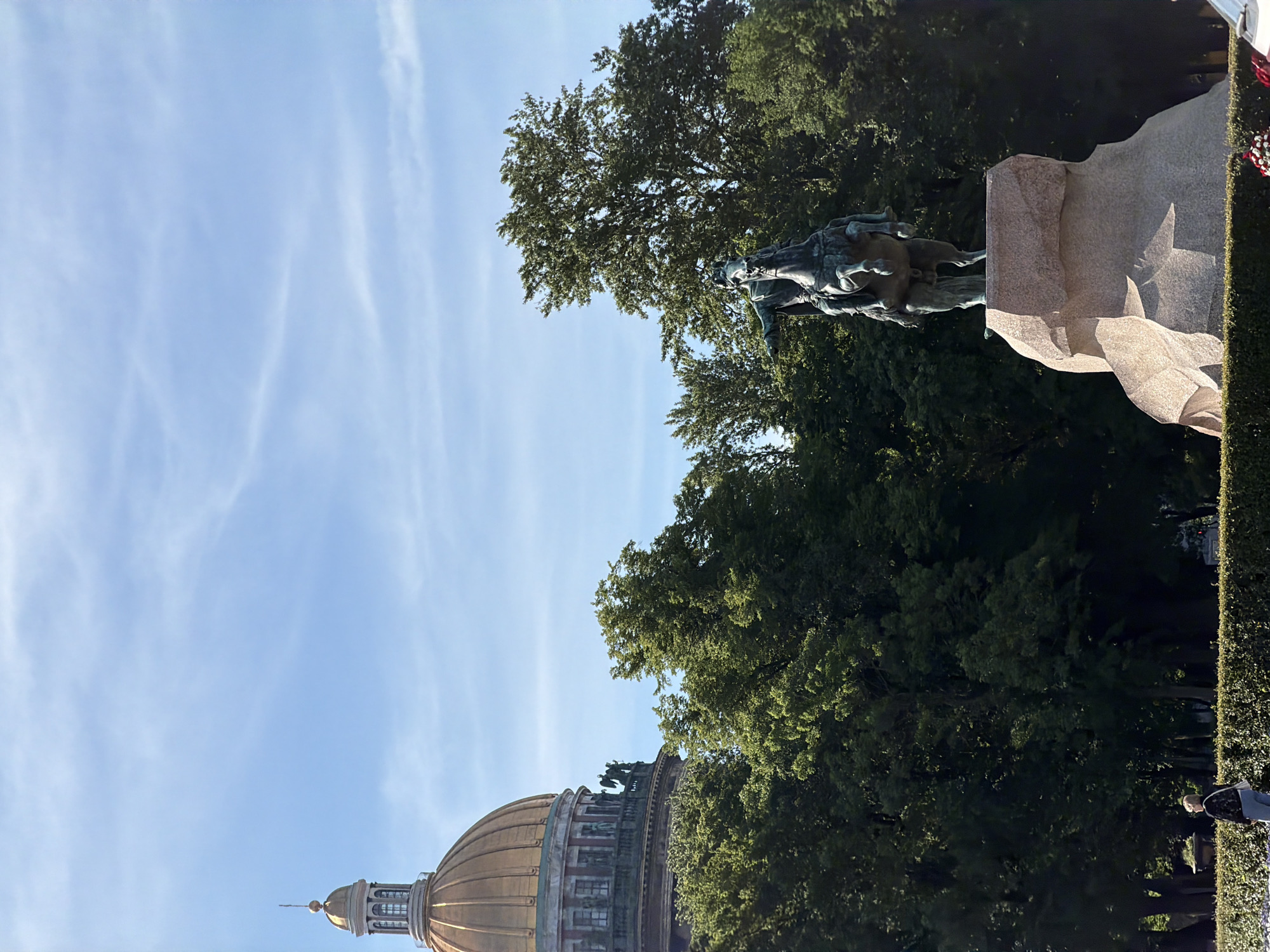 The image shows a full shot of the Bronze Horseman, a statue of Peter the Great on horseback, located in Senate Square in Saint Petersburg, Russia. The statue is made of bronze and stands on a large granite pedestal. It is a bright, sunny day with a clear blue sky and some wispy clouds. In the background, a portion of the dome of St. Isaac's Cathedral is visible, along with lush green trees. In the foreground, a person is visible walking in front of a manicured hedge.