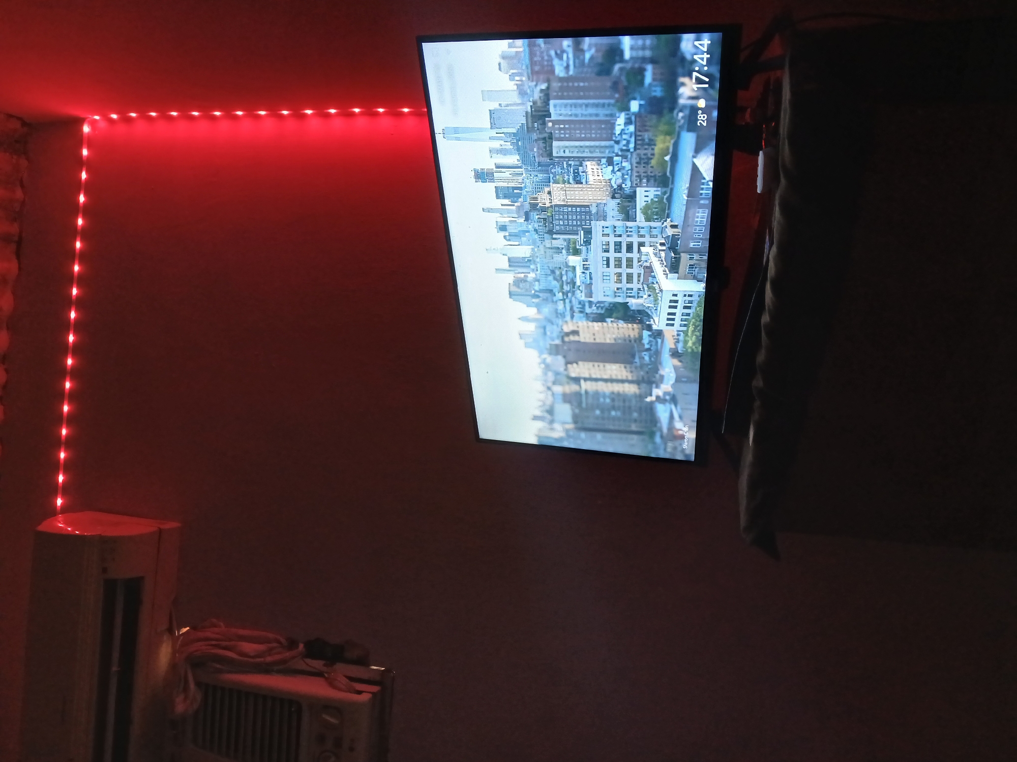 The image shows a dimly lit room with red LED strip lights lining the top corner of the wall. A television screen is mounted on the wall, displaying an urban cityscape. The time "17:44" and temperature "28°" are visible in the upper right corner of the screen. In the lower left corner of the image, there is an air conditioning unit. Part of another smaller appliance is visible next to the AC unit. The wall is a muted reddish-brown color. The scene does not depict the city of Juan Griego, Venezuela, but an urban environment elsewhere. There are no people visible.