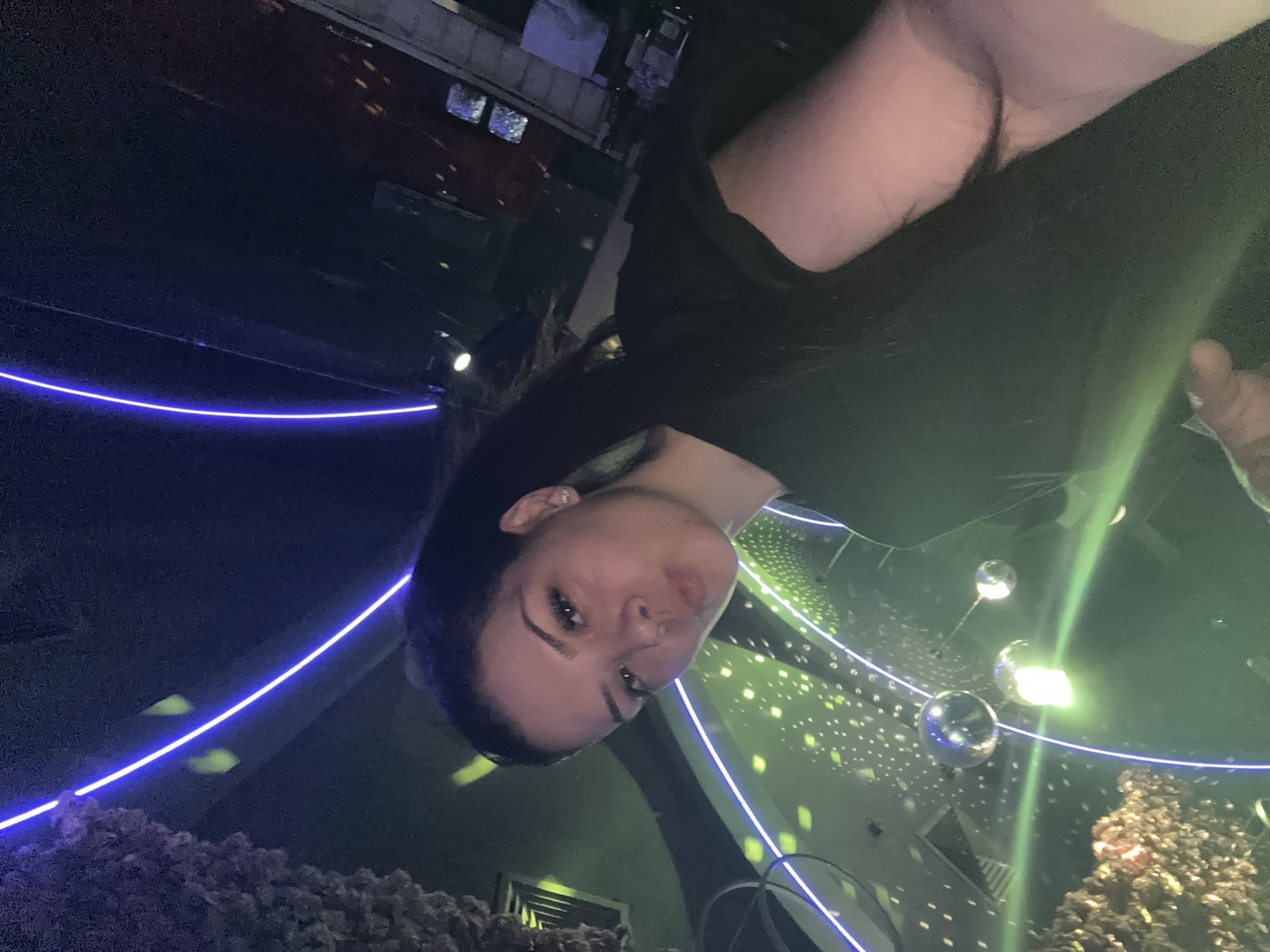 A young woman is captured in a selfie with a disco ball and neon lights in the background, suggesting a club or party setting. The woman is wearing a black shirt and has her hair pulled back. Her expression is playful, with her lips puckered. The environment is dimly lit, with green and purple lights casting an atmospheric glow. Disco balls are visible hanging from the ceiling, reflecting the lights. The image is likely taken in a city like Sliven, Bulgaria, where such entertainment venues are common. The overall impression is one of a fun and vibrant night out.