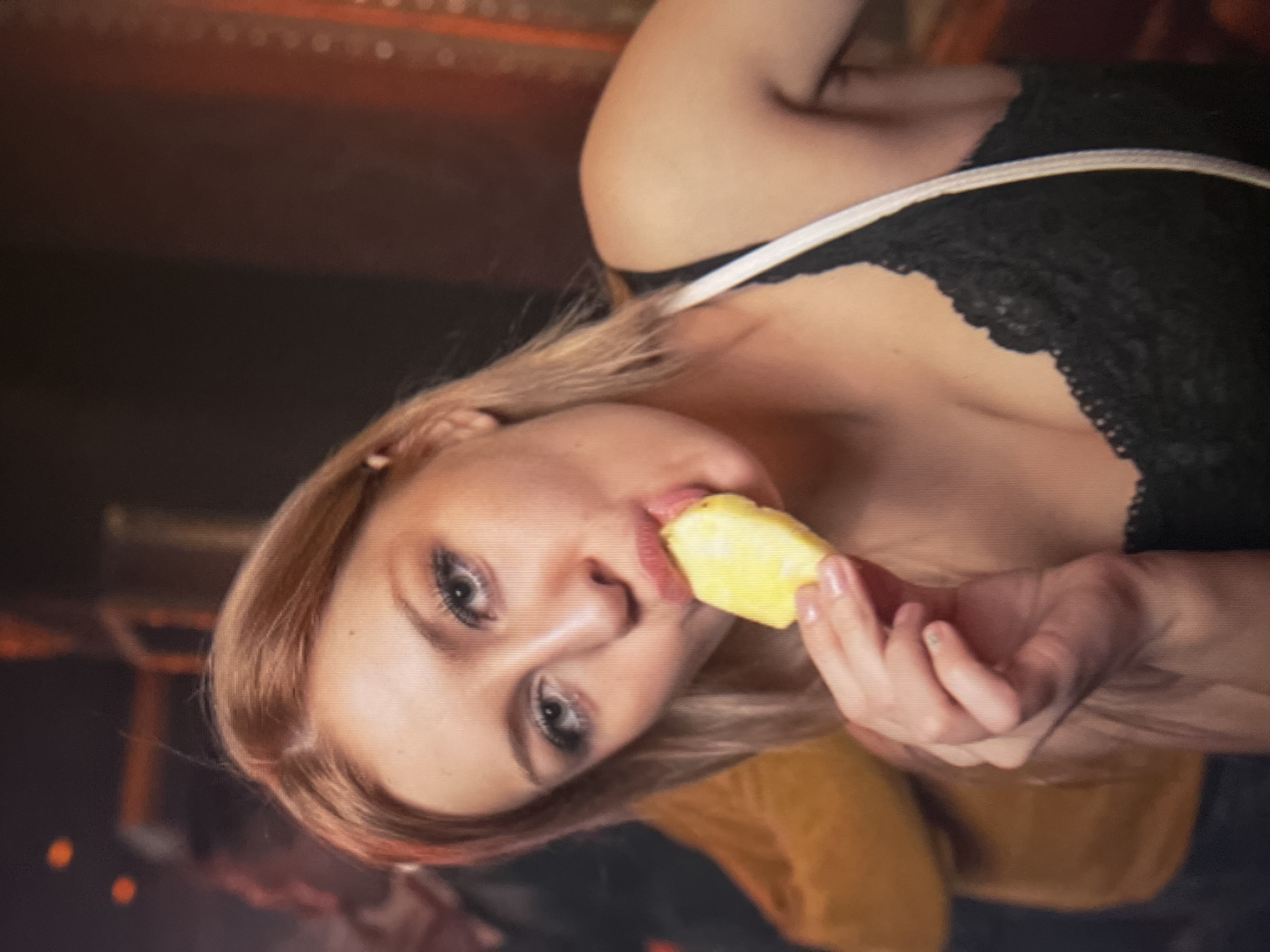 The image shows a close-up of a young woman with blonde hair and light-colored eyes, who is holding and eating a slice of pineapple. She is wearing a black lace top. The background is blurred and appears to be an indoor setting, possibly a bar or club, with some ambient lighting. There are no visible landmarks or elements that would indicate the specific location of Qarağandı city administration, Kazakhstan.