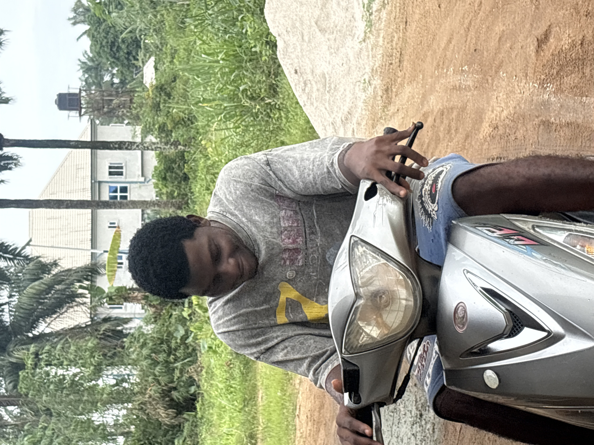 A person with dark skin and short, curly black hair is seated on a silver motorcycle. The individual is wearing a gray long-sleeved shirt with a yellow graphic and blue shorts. The motorcycle's headlight is visible. Behind the subject, a two-story house with a water tower is visible. Lush green vegetation surrounds the building. The ground appears to be a mix of dirt and grass.
The location is presumed to be in Ikot Ekpene, Nigeria.