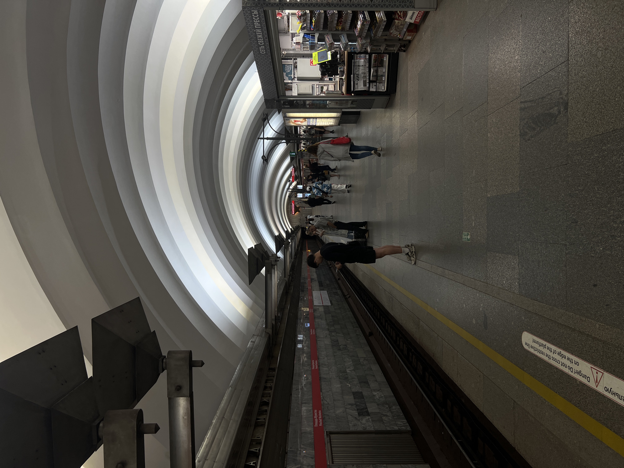 The image depicts the interior of a subway station in Saint Petersburg, Russia, specifically the Ploshchad Muzhestva station. The station features a distinctive arched ceiling with alternating light and dark grey stripes, creating a visually striking effect. The platform is made of granite paving stones, and a yellow line runs along the edge, indicating a safety boundary.
Several individuals are present on the platform. In the foreground, a young man in a black t-shirt and shorts stands looking at his phone. Further down the platform, other passengers can be seen walking and waiting for trains. To the right, a kiosk labeled "СЕТЬ СВЕЖЕЙ ПРЕССЫ" (Press News Network) is visible, stocked with newspapers, magazines, and other items. The walls of the station are clad in marble, and overhead lighting fixtures are mounted on metal structures. The train tracks are visible to the left, with a train partially visible in the distance. The overall atmosphere is one of an active, albeit somewhat dimly lit, urban transit environment.