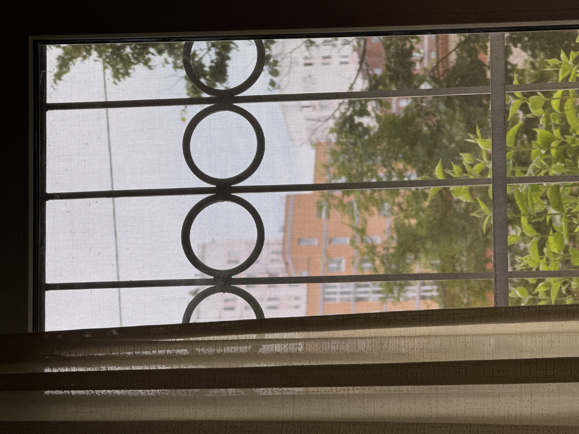 An exterior view of a residential building is visible through a window with a metal security grate and screen. A beige curtain is partially drawn to the left of the frame. The window is divided by vertical bars with decorative circular elements. Beyond the grate, a multi-story building with orange and white facades and numerous windows is partially obscured by green foliage. The sky appears overcast. The scene suggests a view from a window in an urban environment, possibly in Khimki, Russia, given the architectural style and general impression. There are no people or animals present in the image.