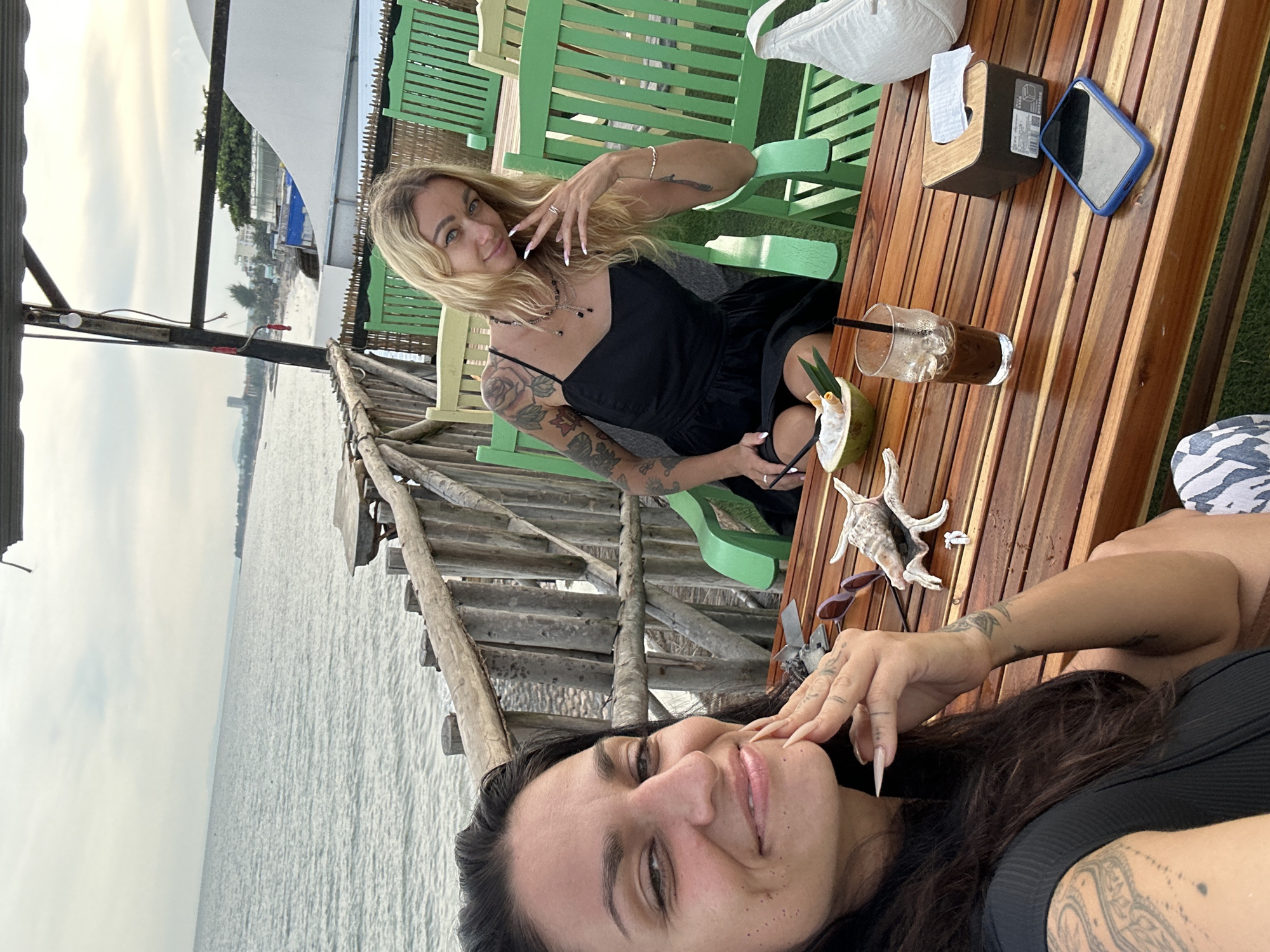 The media file features two women in an outdoor setting, likely a beachside cafe or restaurant in Phu Thuy Ward, Vietnam. One woman, with blonde hair and tattoos, is seated on a green chair at a wooden table. She is wearing a black dress and jewelry, and appears to be posing for the photo. She has a drink in front of her, and a coconut with a straw. The second woman, with dark hair and tattoos, is in the foreground, taking the selfie. She is wearing a black top and is smiling at the camera.
The scene is set by the ocean, with calm water visible in the background. The sky is overcast, suggesting it might be morning or late afternoon. The wooden table has a tissue dispenser, a phone, and a drink, all indicating a relaxed atmosphere where people might be enjoying a meal or drinks. The overall mood appears to be casual and social. The wooden structures and furniture, along with the beachside view, suggest a tropical or coastal environment.