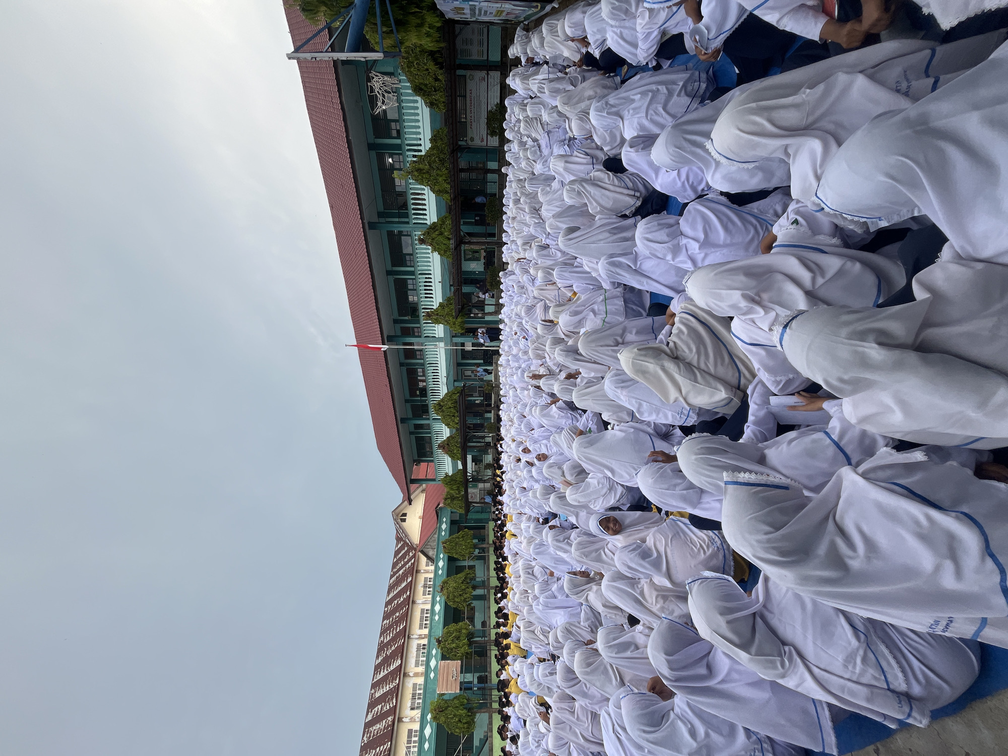 This image depicts a large gathering of students, predominantly female, seated on the ground outdoors. The majority of the students are wearing white hijabs and uniforms, creating a visually uniform crowd. They appear to be attentive, with many looking towards a central point or area not fully visible in the frame. In the background, there is a building with a red-tiled roof and teal-colored trim, which appears to be a school or institutional structure. A flag is visible on a flagpole in front of the building, suggesting an official event or assembly. Trees and some basketball equipment are also present in the background. The lighting suggests an overcast day. The presence of the school building and the large number of students in uniform strongly suggests an educational setting, possibly during a school event or assembly. The image context indicates this scene is located in Lhokseumawe, Indonesia.