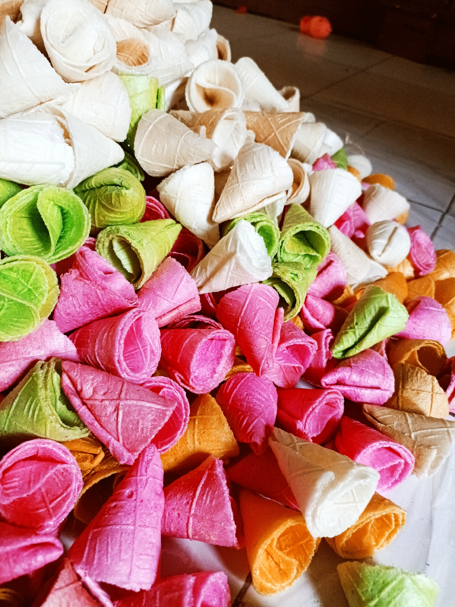 A pile of colorful wafer cones is displayed on a white surface, possibly a table or a floor. The cones are of various sizes and shapes, some are fully formed into cones, while others appear to be rolled up. The colors include pink, green, orange, white, and beige. Some of the cones have a waffle-like pattern imprinted on them. In the background, a tiled floor is visible, and a blurred orange object is present. The image appears to be taken in the city of Jatitengah, Indonesia, suggesting this could be a local delicacy or confectionery.