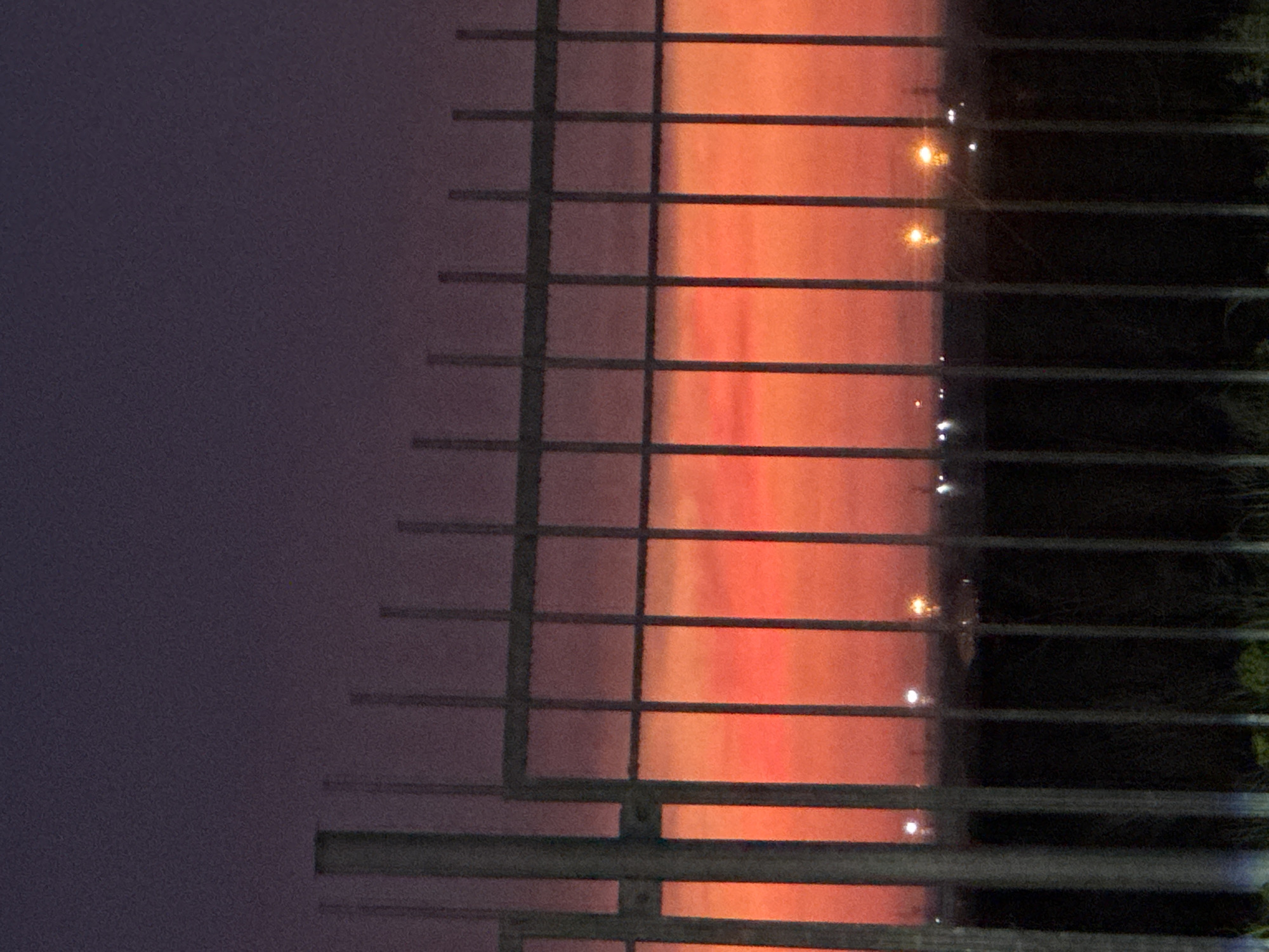 A high-angle outdoor photograph captures a metal security fence silhouetted against a dramatic twilight sky, likely sunrise or sunset. The sky transitions from a deep, desaturated purplish-gray at the top of the frame to a vibrant, intense orange-red hue near the horizon. The fence, composed of numerous evenly spaced vertical bars and at least five visible horizontal rails, forms a grid pattern across the central and lower portions of the image. A thicker, primary vertical post frames the far-left edge. Below the vivid horizon, a dark, silhouetted landscape or ground mass is visible, dotted with multiple small, intensely bright, out-of-focus artificial light sources, suggesting distant urban or industrial infrastructure. The immediate foreground below the fence is a dark, blurred area, possibly containing sparse vegetation. The entire image exhibits noticeable visual noise, characteristic of low-light photography. No people, subjects, or explicit actions are present. The specific location within Unknown, Unknown, cannot be determined from the visual information.