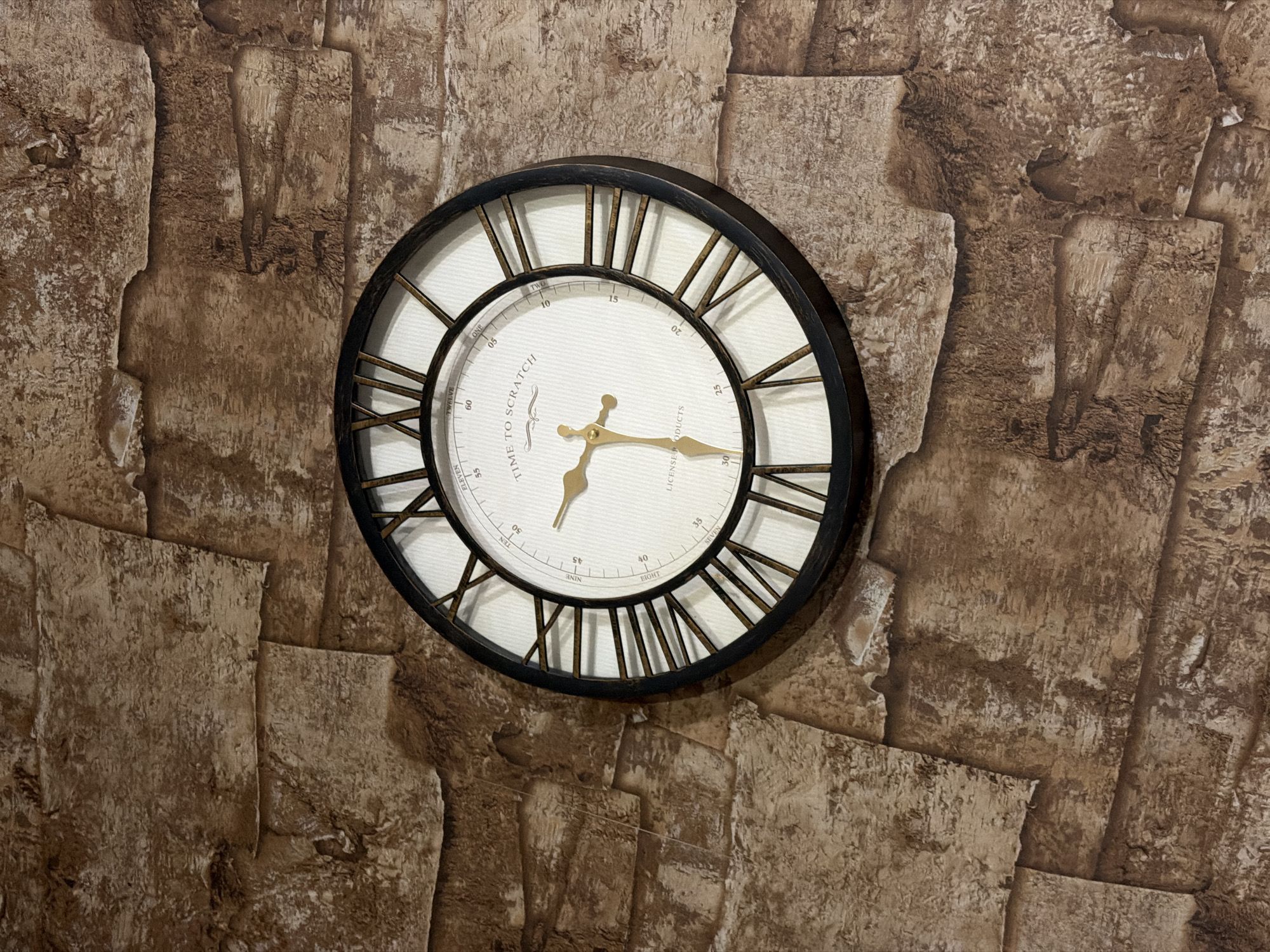 A wall clock is affixed to a textured brown wall. The clock has a black circular frame, Roman numeral markings, and gold-colored hands indicating the time. The clock's face includes the phrase "TIME TO SCRATCH." The wall's texture resembles stacked stones.