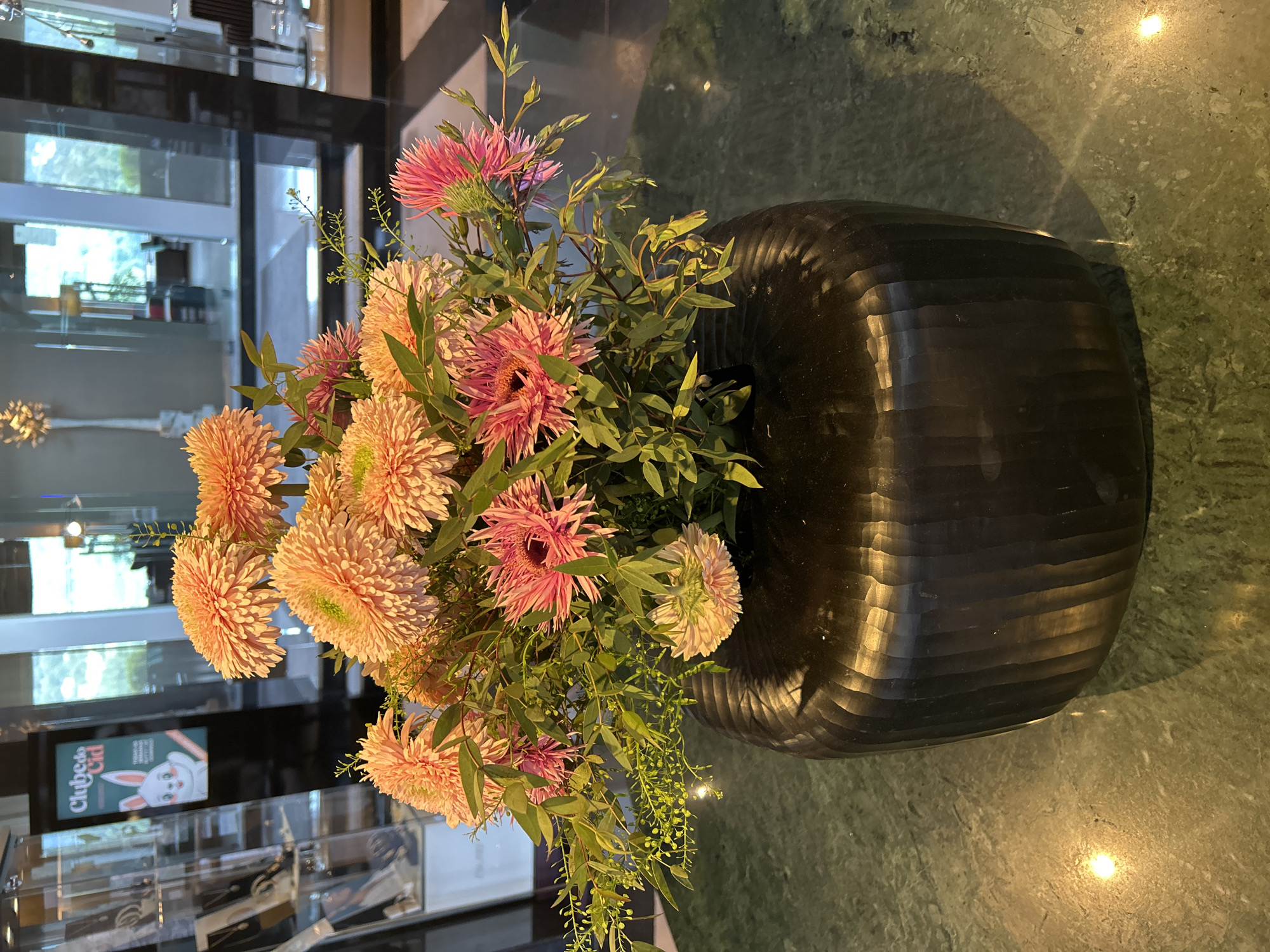 A close-up, eye-level shot focuses on a large bouquet of vibrant flowers in a dark, ribbed vase. The flowers are a mix of peach and pink hues, with some delicate green foliage and tiny buds interspersed throughout. The vase, made of a dark, textured material, sits on a polished green countertop with a mottled pattern. The background is softly blurred, revealing glimpses of an indoor environment with glass partitions, reflections, and what appears to be a retail display case on the left. The lighting casts subtle shadows and highlights on the vase and countertop, suggesting an ambient indoor setting.
