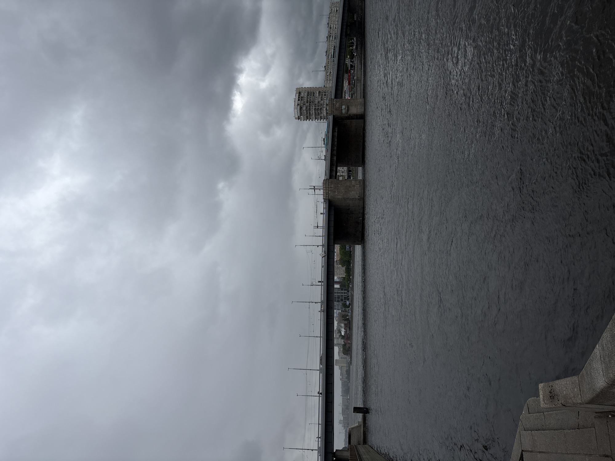 The image depicts a dark and overcast scene in Saint Petersburg, Russia. A river fills the right half of the frame, its surface rippled by wind. The left half is dominated by a heavy, grey sky. A bridge stretches horizontally across the middle ground, appearing as a long, dark line punctuated by support pillars and light posts. A few buildings are visible along the horizon line on the far side of the river. A stone embankment is visible in the lower right corner of the frame. No people are visible.