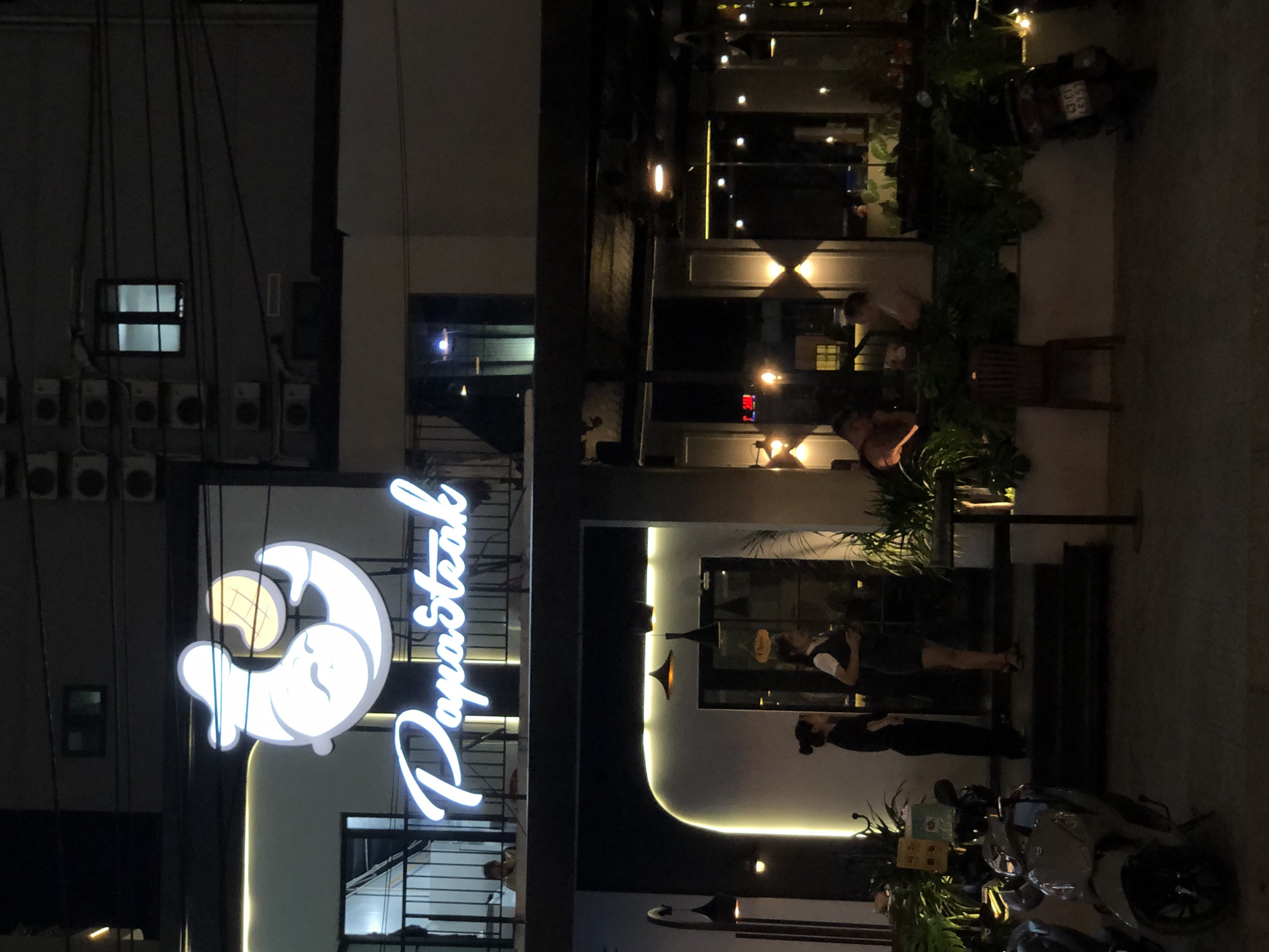 This is a nighttime outdoor photograph of a restaurant in Da Nang, Vietnam, identified by the sign "Papasteak" in illuminated white script. The restaurant features a modern, multi-level facade with dark balconies and glowing white accent lighting. On the upper level, a cartoonish white logo of a shrimp holding a potato slice is visible, also illuminated. Below, on the ground level, a wide entrance with a curved, illuminated opening leads into the establishment. Two women in uniform, presumably staff, are standing near the entrance. A motorcycle is parked in the foreground on the left, and another is partially visible on the right. Lush green plants surround the entrance and are integrated into the building's design, creating a welcoming ambiance. The street in front of the restaurant appears to be paved.