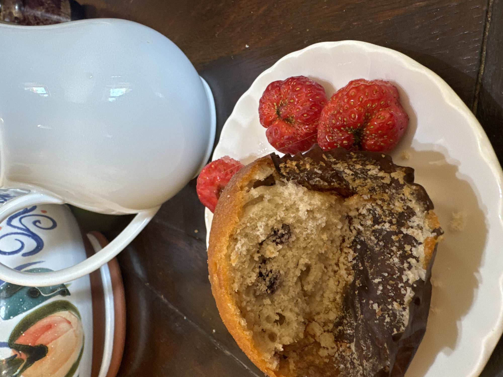A partially eaten muffin with chocolate frosting and blueberries is on a white plate with three strawberries. The plate is on a dark wooden table. In the background, a white ceramic pitcher and a colorful ceramic bowl are visible. The scene is indoors, with no specific location in Waterford, Ireland identified.