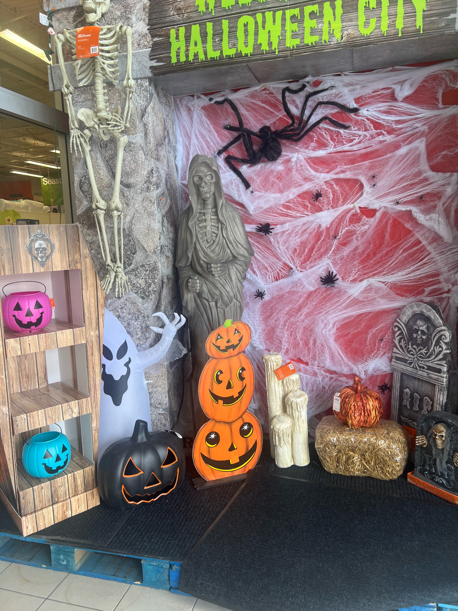 The image depicts a Halloween display, likely set up outside a retail store, possibly in the city of Toronto, Canada. The display is arranged on a wooden pallet, with a black mat in front of it.
Dominating the scene is a large, nearly life-sized skeleton hanging from the top left. To its right, a tombstone with a skull and the word "RIP" is visible. Behind these elements, a red backdrop is covered in white artificial cobwebs, populated by several black plastic spiders of varying sizes, and one very large spider hanging prominently. A figure resembling the Grim Reaper, draped in a hooded robe, stands to the right of the skeleton.
In the foreground, a wooden shelf unit displays two jack-o'-lantern buckets: one pink on the top shelf and one turquoise on the second shelf. To the right of the shelf unit, an inflatable white ghost with a black face stands partially obscured by the Grim Reaper figure. A stack of three orange pumpkins, also carved as jack-o'-lanterns, is positioned to the right of the ghost.
Further to the right, a bale of straw is topped with a decorative orange pumpkin. Beside the straw bale, a set of three large, cream-colored artificial candles stands upright. To the far right, a small tombstone-like figurine depicts a skeleton emerging from a grave.
A wooden sign above the cobwebbed backdrop reads "HALLOWEEN CITY" in dripping green letters. The environment appears to be an outdoor or semi-outdoor retail space, with a glimpse of a store interior visible in the background on the left, including shelving and signs.