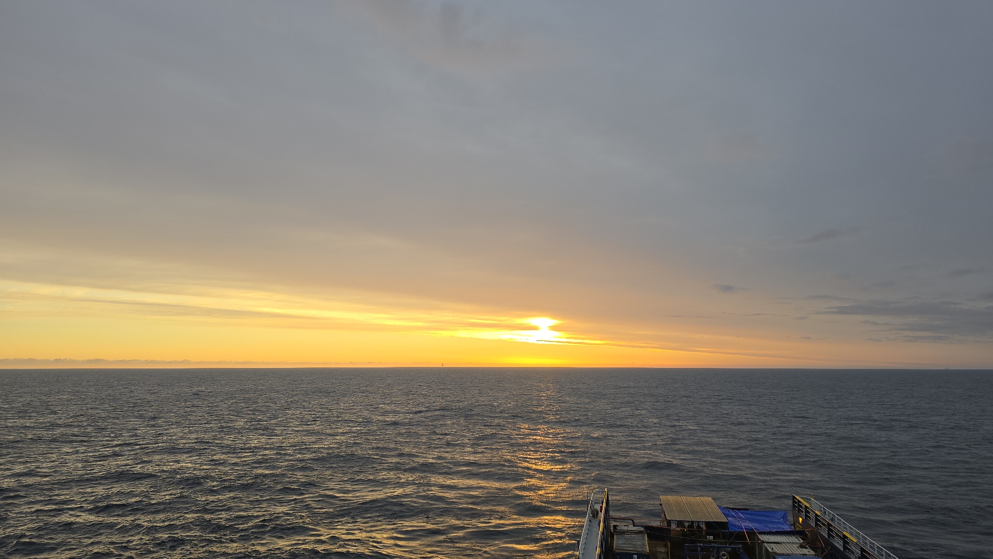 The image depicts a sunset over the ocean, likely taken from a boat or ship. The sky transitions from a pale gray at the top to a vibrant yellow and orange near the horizon, where the sun is setting. The sun's reflection creates a bright, shimmering path on the water's surface. The ocean itself is a dark, slightly choppy expanse. In the foreground, parts of the boat or ship are visible, including railing, containers, and equipment. The location appears to be open ocean.