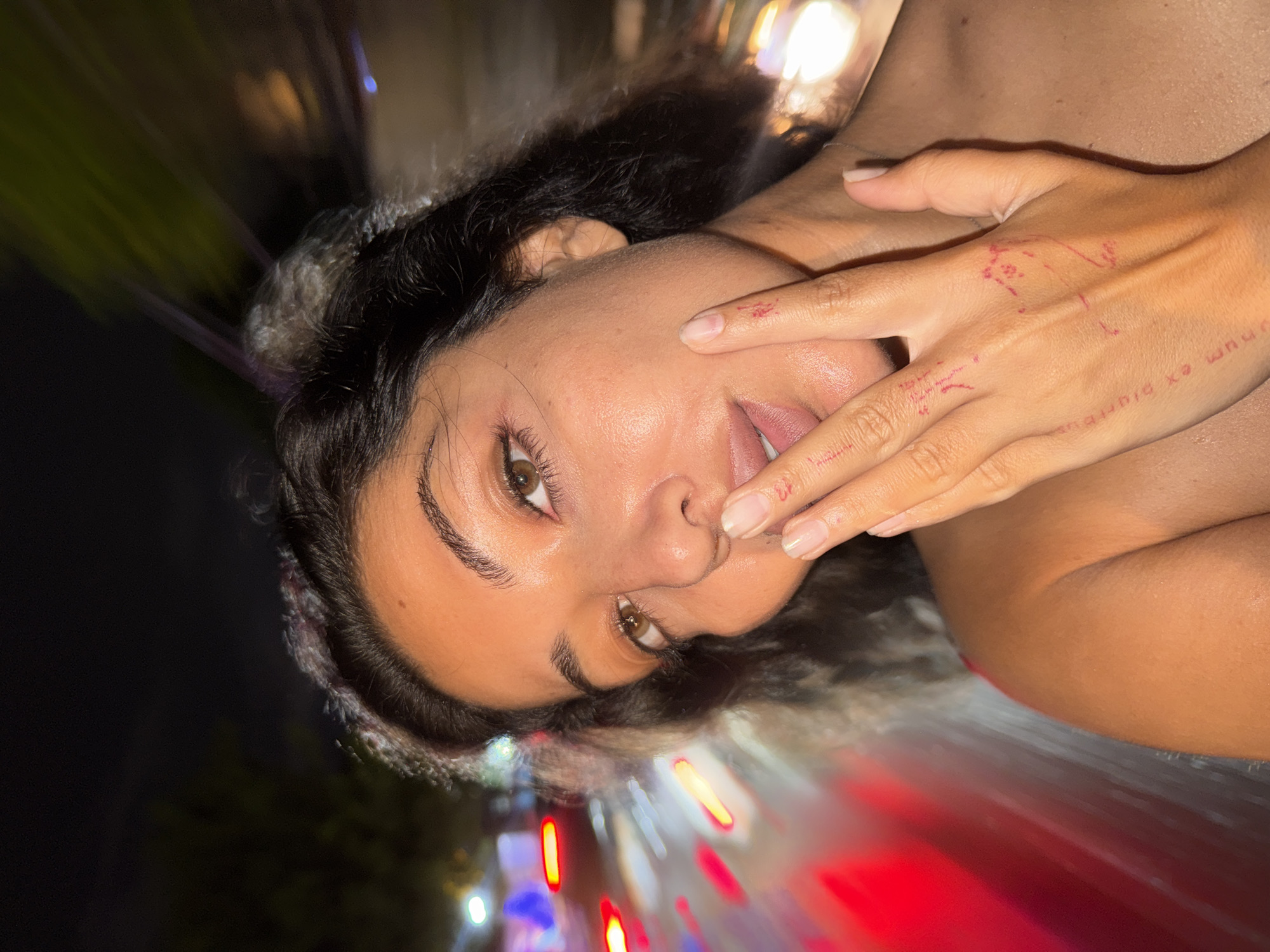 A young woman is depicted in a close-up shot, positioned at an oblique angle, against a background suggestive of an urban night scene. Her face is prominently displayed, revealing brown eyes, dark hair, and clear skin. One hand is raised to her face, with faint markings or inscriptions visible on the hand. The blurred background features streaks of red and white light, suggesting motion or a dynamic environment. The North Kuta, Indonesia, location is not discernible from the visual information provided.