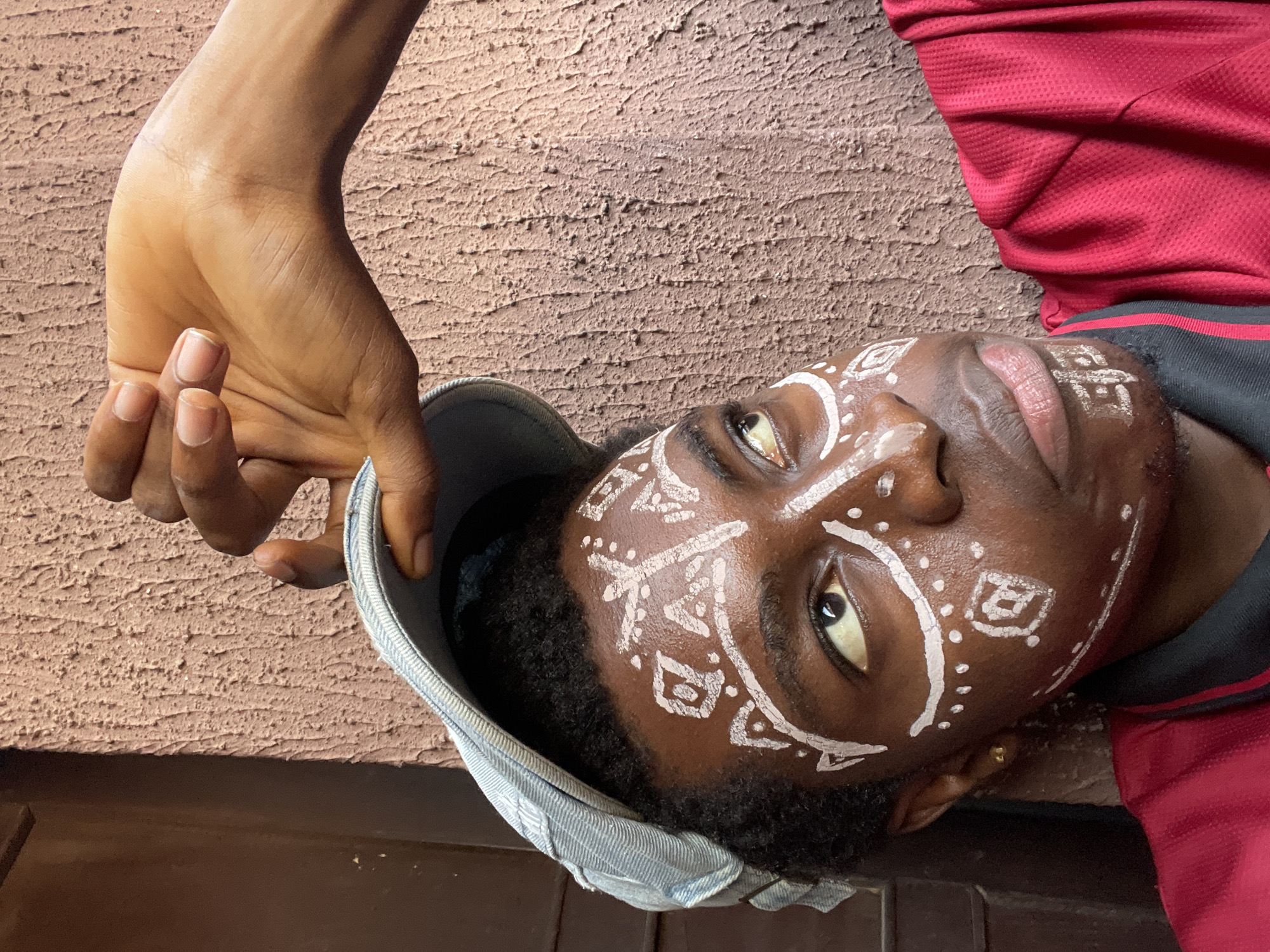 A close-up, medium-lit shot depicts a person, possibly in Nkpor, Nigeria. The person, positioned in the lower-center of the frame, has dark skin and distinct white face paint in tribal patterns. They wear a light blue denim cap held in place by a light-skinned hand, the fingers gently curved. The person is wearing a red and black shirt. The background features a textured, light brown or beige wall. The framing cuts off part of the person and the wall.