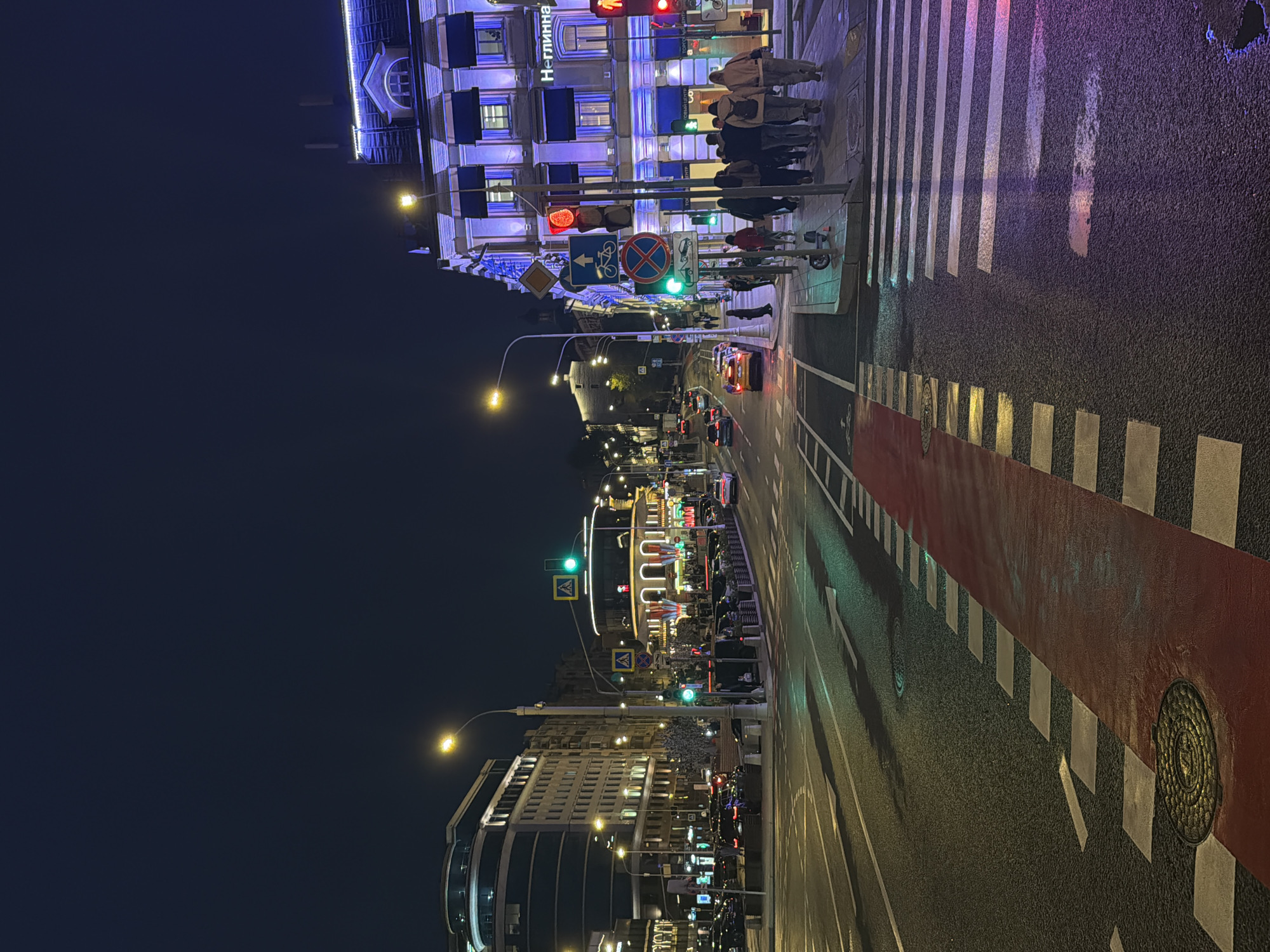The image shows a nighttime street scene in Moscow, Russia. The view is along a street lined with buildings, illuminated by streetlights and building lights. In the foreground is a pedestrian crosswalk with people crossing. A bicycle lane, marked in red, runs along the street. Traffic signals are visible, some showing green lights. Cars are driving on the street. The buildings vary in architectural style, with one prominently featuring a sign in Cyrillic script ("Неглинна"). The sky is dark, suggesting a night scene. The street appears wet, reflecting the lights.