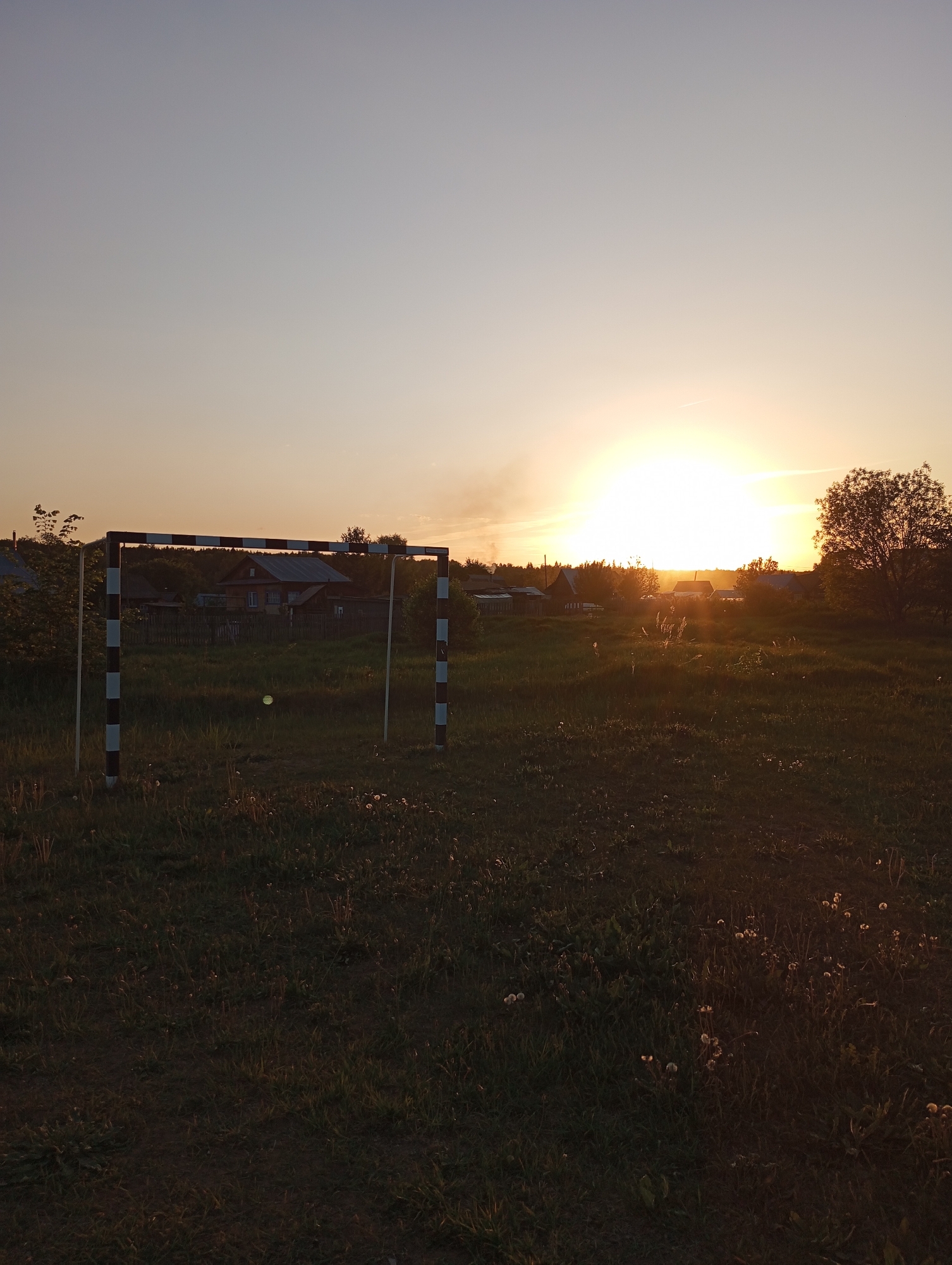 The image shows a field at sunset near the city of Буренка, Russia. A black and white striped soccer goal stands in the foreground. Beyond the goal, a cluster of buildings is visible, likely houses or other structures within the city or a nearby village. The sun is setting on the horizon, creating a bright glow in the sky with scattered smoke, suggesting a possible fire or distant industrial activity. The field is covered with grass and small plants, some of which appear to be dandelions.