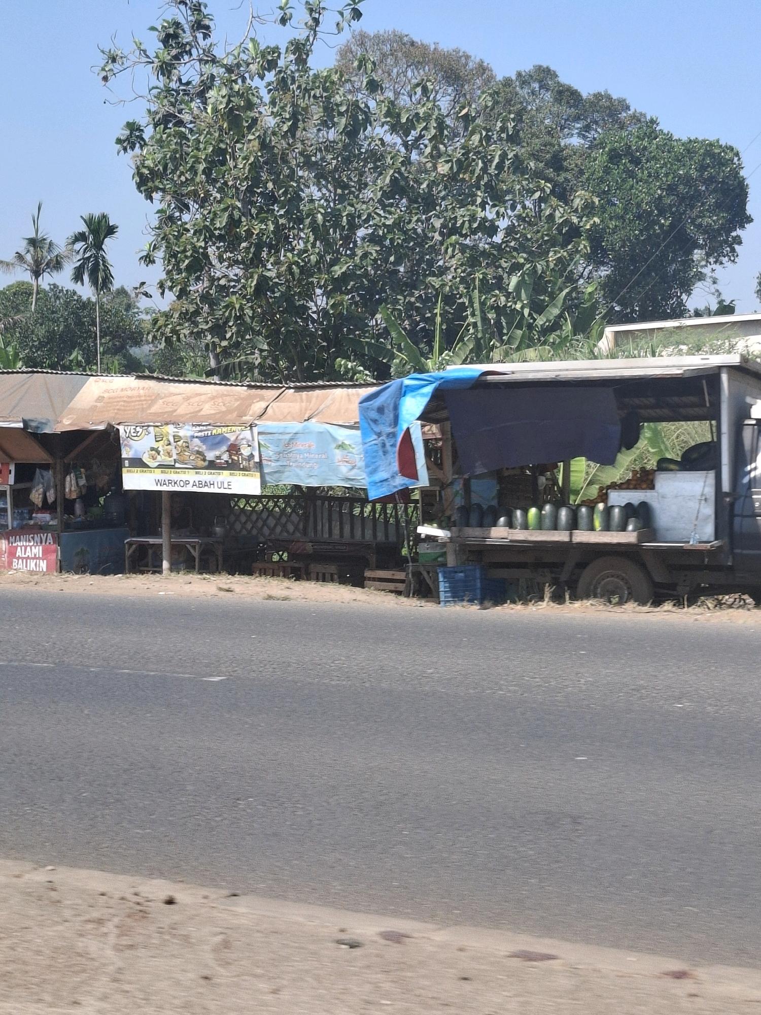 The image shows a roadside scene in Babakan, Indonesia. A small roadside stall with a corrugated metal roof is visible, featuring various items for sale, including what appear to be snacks or packaged goods. A banner advertising "WARKOP ABAH ULE" is prominently displayed, suggesting a coffee shop or food stall. To the right, a truck serves as a makeshift market stall, laden with watermelons. The truck is covered with tarps, and the watermelons are arranged in crates on the truck bed. In the background, lush green trees and vegetation are abundant, typical of a rural or semi-rural Indonesian landscape. The road in the foreground appears to be asphalt, with a sandy shoulder. The sky is clear and blue.
