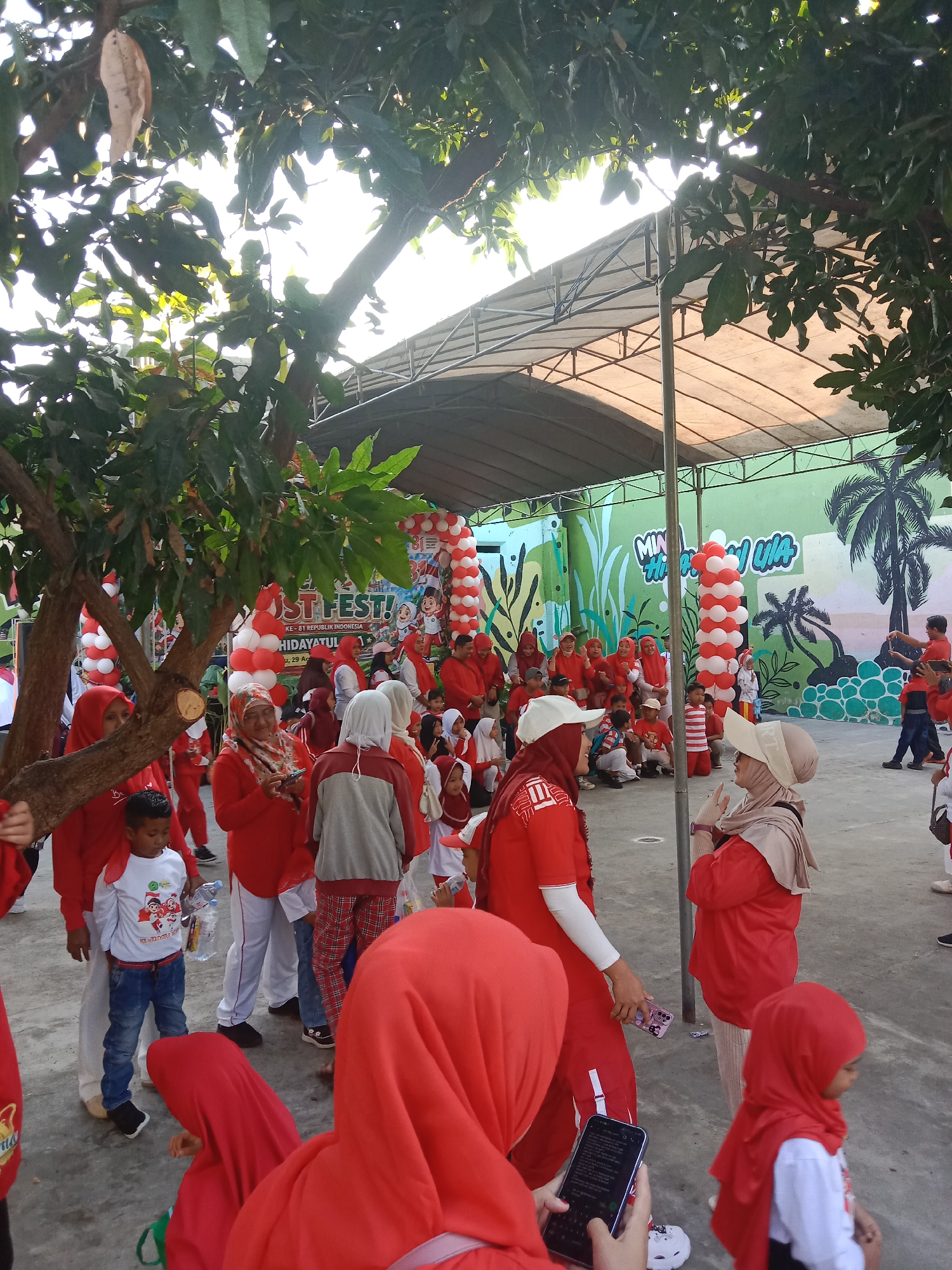 The image depicts an outdoor gathering, likely a celebration or festival, on a paved ground area. A canopy structure is visible in the background, supported by poles. The scene is decorated with red and white balloons, arranged in columns. A colorful mural adorns the wall behind the canopy, featuring stylized plants and palm trees. A banner with text, partially obscured by balloons and foliage, reads "ST FEST!" and "KE-81 REPUBLIK INDONESIA".
A significant number of people, mostly dressed in red attire and headscarves, are present. Among them are adults and children. Some individuals are standing and interacting, while others are seated or in motion. In the foreground, a young boy in a white t-shirt and blue jeans holds a water bottle. The surrounding environment includes large trees with green leaves, casting shadows on the ground. The lighting suggests it is daytime, possibly late afternoon, with sunlight filtering through the foliage. The overall atmosphere appears to be festive and communal.