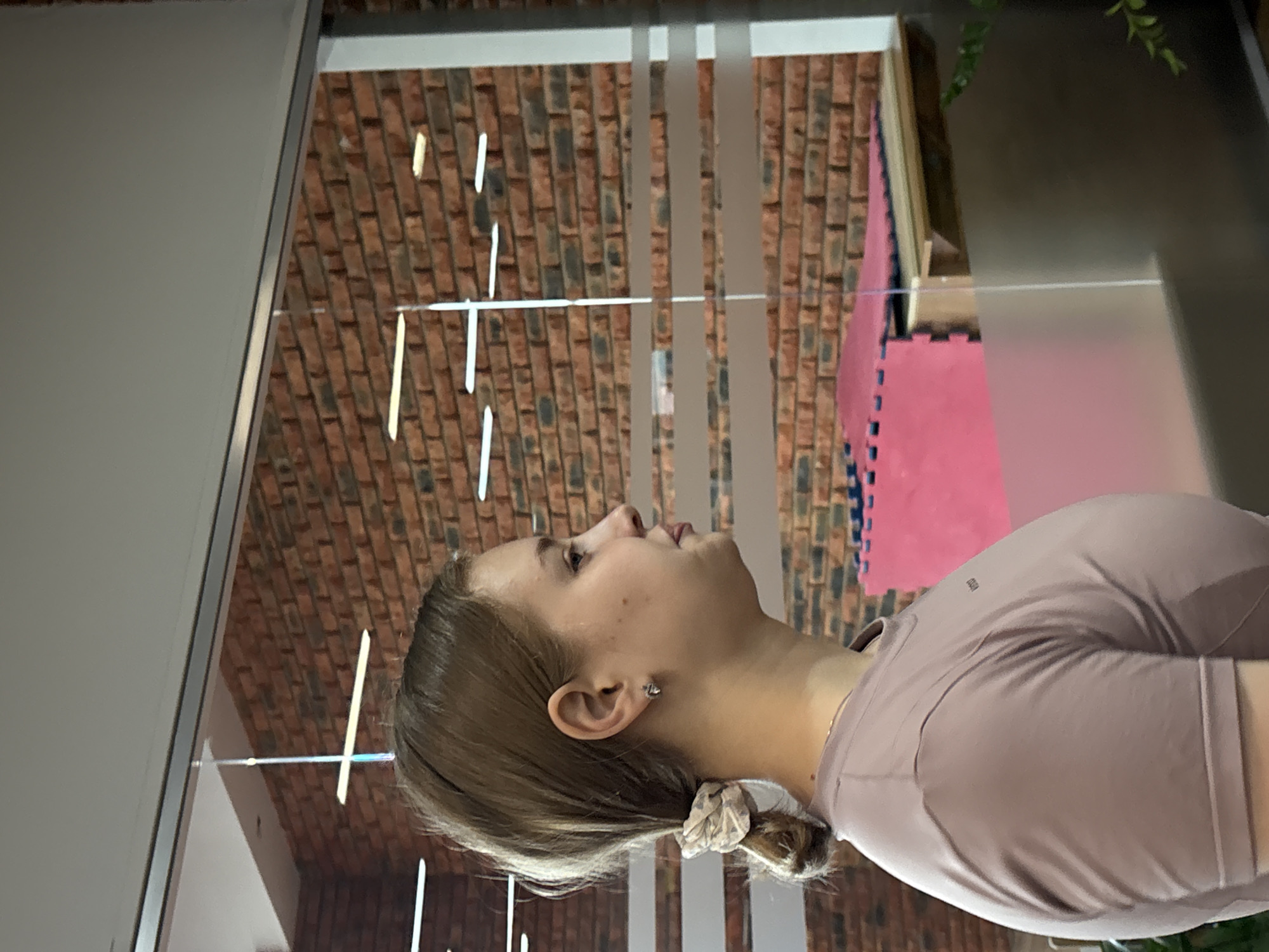 A young woman is depicted in a profile, chest-up shot, looking towards the upper left of the frame. Her light brown hair is pulled back, with a light-colored scrunchie visible at the nape of her neck. She wears a plain, light mauve or dusty pink long-sleeved athletic top, with a small dark logo (possibly "lululemon") visible on her upper back. A small, simple earring adorns her visible ear. Her expression is neutral and observant, as if looking at something above or in the distance.
The setting appears to be indoors, likely a modern facility or studio. In the background, a rustic red brick wall is visible through a series of clear glass panels, which are segmented by vertical frosted strips and feature thin horizontal lines. On the floor in the mid-ground, vibrant pink interlocking foam mats are laid out, suggesting a space used for physical activities such as martial arts, gymnastics, or dance. A small amount of green foliage is peeking into the frame from the top right, behind the glass. The lighting suggests daytime. The overall impression is that of a clean and functional indoor environment in Chaikovsky, Russia.