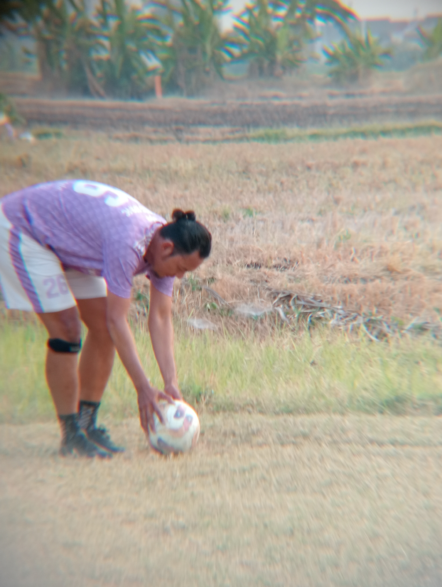 An unidentified male wearing a purple jersey and white shorts with the number "26" visible on the left leg, is bending over a soccer ball on a dry, grassy field. He has his hair tied in a bun. A black knee brace is visible on his left knee, and he is wearing dark socks and athletic shoes. The background shows a blurred landscape with vegetation. The image quality is low, with a noticeable blur and color fringing, making specific details about the location in Kenep, Indonesia difficult to ascertain, but the presence of dry grass and scattered vegetation suggests a rural or semi-rural outdoor environment.
