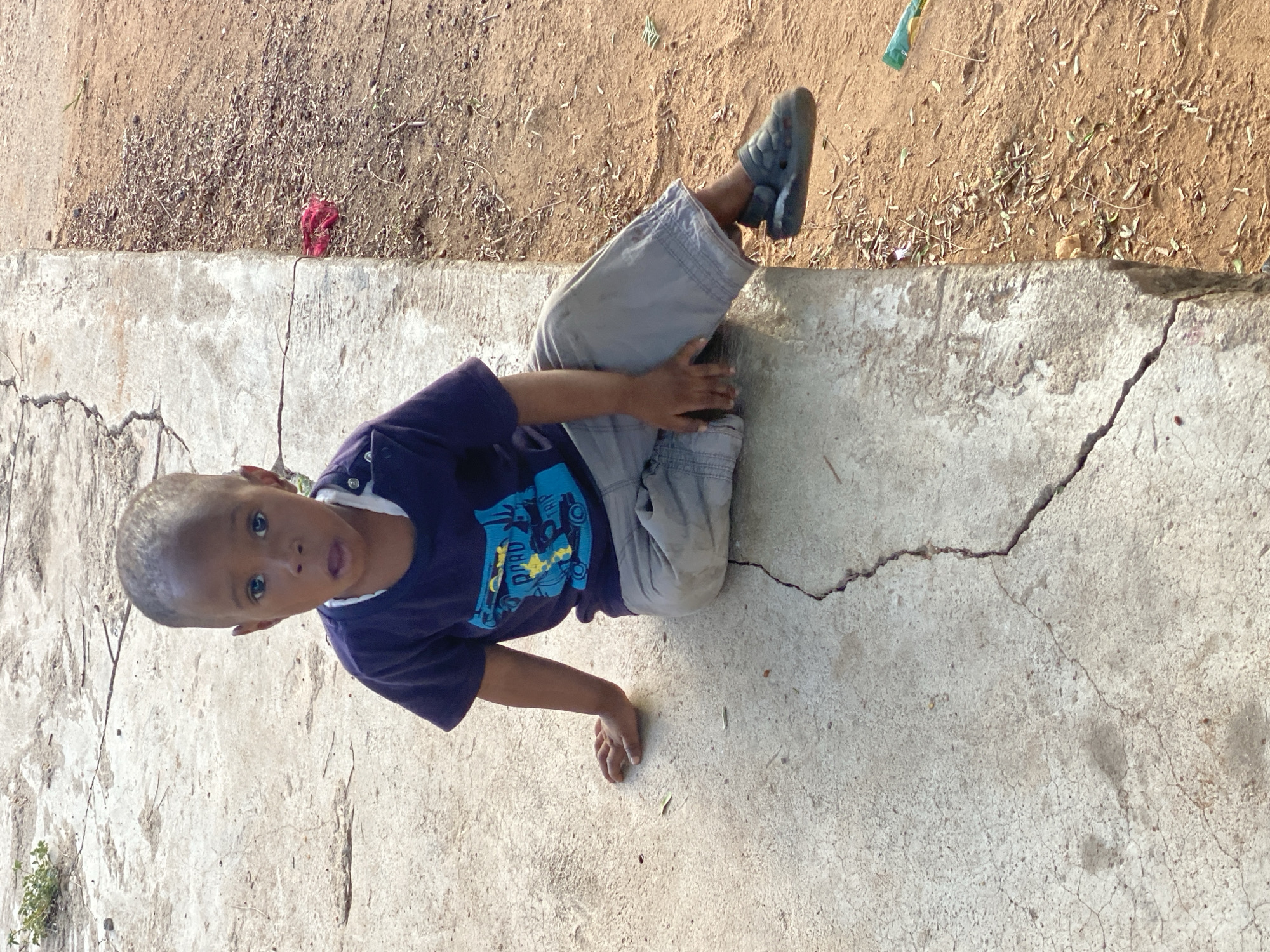 A young child is captured sitting on a concrete surface in an outdoor setting in Adankolo, Nigeria. The child, a boy with short dark hair, is wearing a navy blue short-sleeved shirt with a vibrant light blue and yellow graphic design featuring what appears to be a cartoon vehicle and some stylized, indistinct text. He also wears light grey cargo shorts and dark closed-toe sandals.
The boy is positioned with his legs bent and feet resting on the concrete, looking directly at the camera with wide, curious eyes and a slightly open mouth, conveying an expression of mild surprise or wonder. His left hand rests on his leg, while his right hand is placed flat on the concrete beside him.
The concrete surface he sits on shows signs of age with several cracks. Adjacent to the concrete is a patch of dry, sandy earth, littered with sparse dry leaves, twigs, and a few small green plant shoots. A crumpled green item and a small piece of red fabric are also visible on the ground. The scene is brightly illuminated by natural daylight, indicating clear, sunny weather.