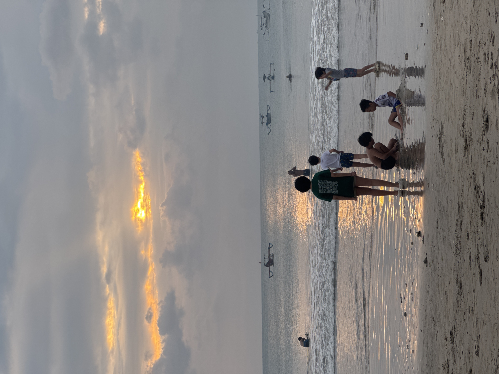 A group of people, primarily children, are at the beach in Kuta Selatan, Indonesia. The scene depicts a sandy shore with gentle waves lapping at the edge. Several individuals are scattered across the beach, some interacting with the water, while others appear to be sitting or playing. One person, wearing a green shirt, is standing and looking towards the water. Further out in the ocean, several small boats, likely traditional fishing vessels, are visible.
The time of day appears to be either sunrise or sunset, as indicated by the warm, golden light reflecting off the water and illuminating the clouds. The sky is partly cloudy, with dramatic streaks of light breaking through. The overall mood of the image seems peaceful and relaxed, suggesting a casual outing at the beach. There is no visible text in the image.