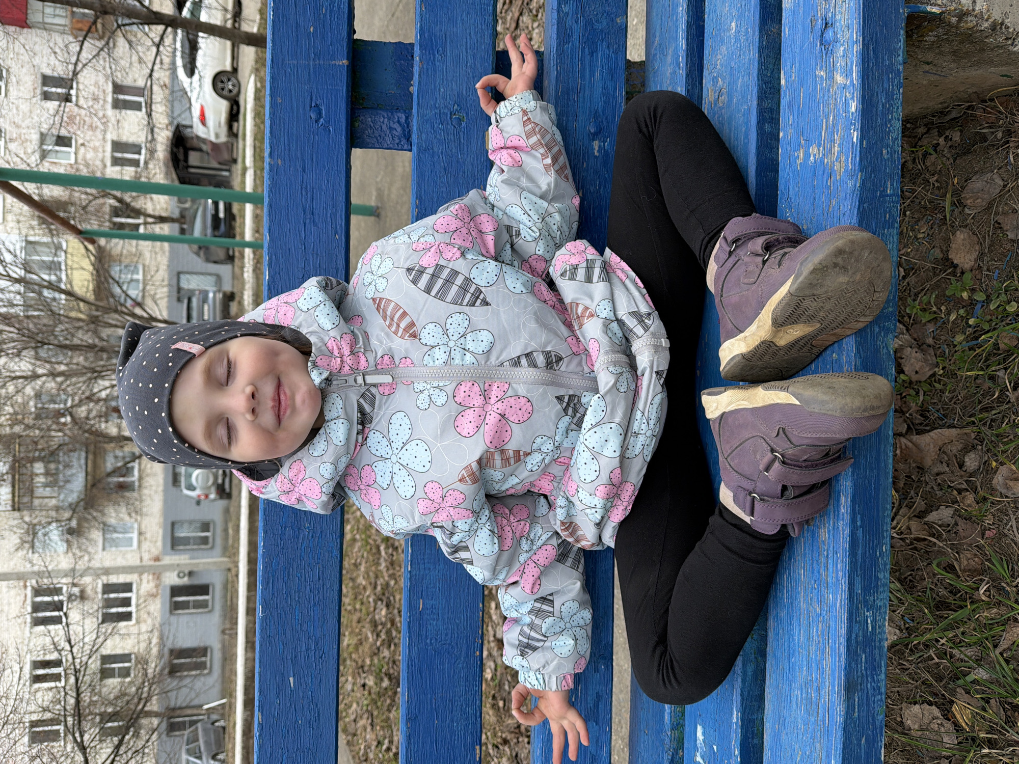 A young child is resting on a blue wooden bench in what appears to be a public park or play area. The child, likely a girl, is lying on her back with her eyes closed, suggesting she might be asleep or resting. She is dressed in black leggings, purple sneakers, and a patterned light gray hooded jacket with pink and light blue floral designs, topped with a gray polka dot hat. The bench is made of parallel wooden planks painted bright blue. The soles of her shoes are dirty. The background shows urban elements including buildings, trees without leaves, and parked cars. Although I can not see the city of Votkinsk, Russia, it can be implied from the information provided.