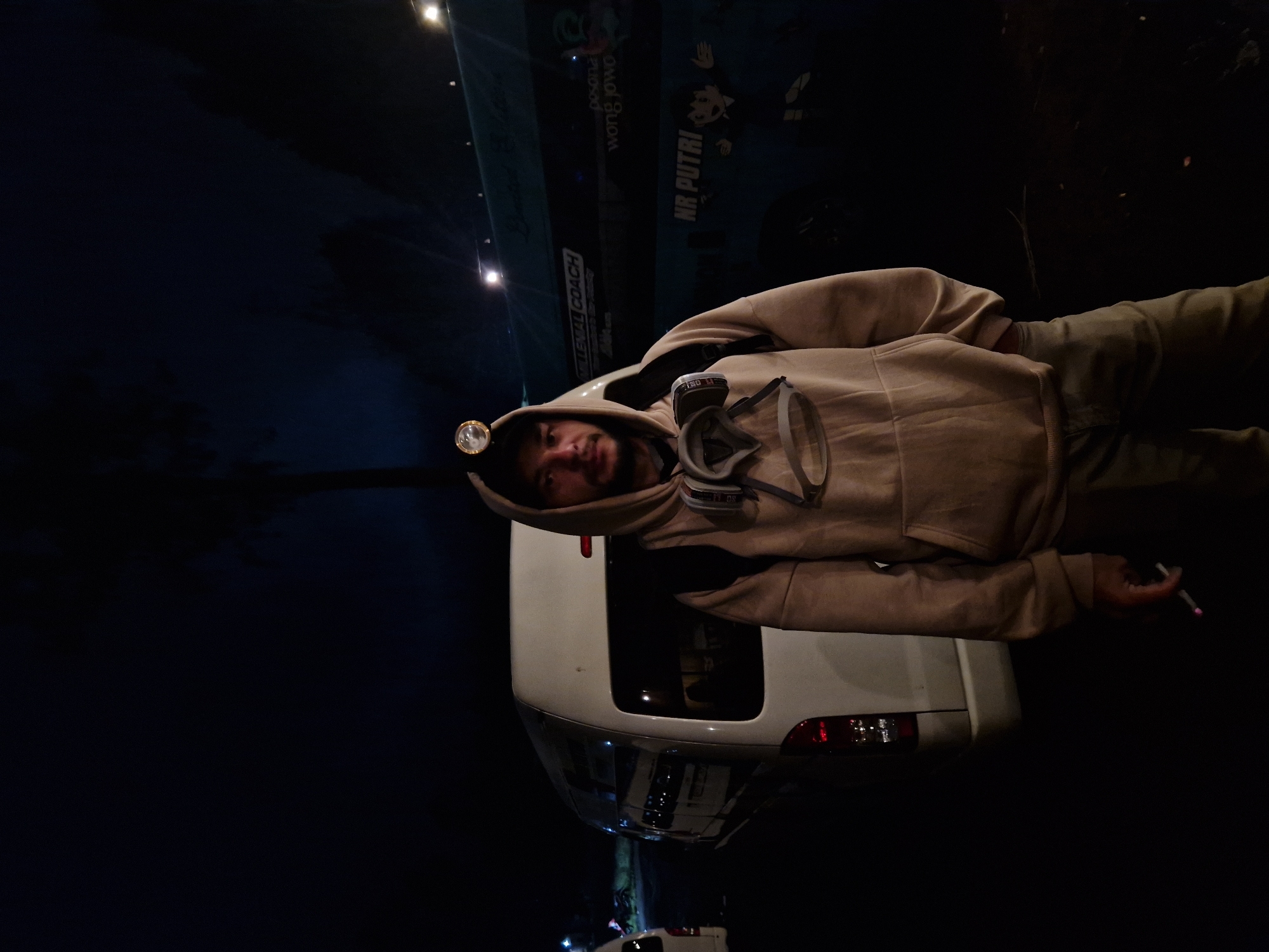 The image shows a man in a tan hoodie and pants, standing next to a white van at night. He is wearing a headlamp and a respirator or gas mask around his neck. He holds a cigarette in his right hand. The background includes a sign with "NR. PUTRI" and Indonesian text, and streetlights in the distance. The location is in the city of Unknown, Indonesia.