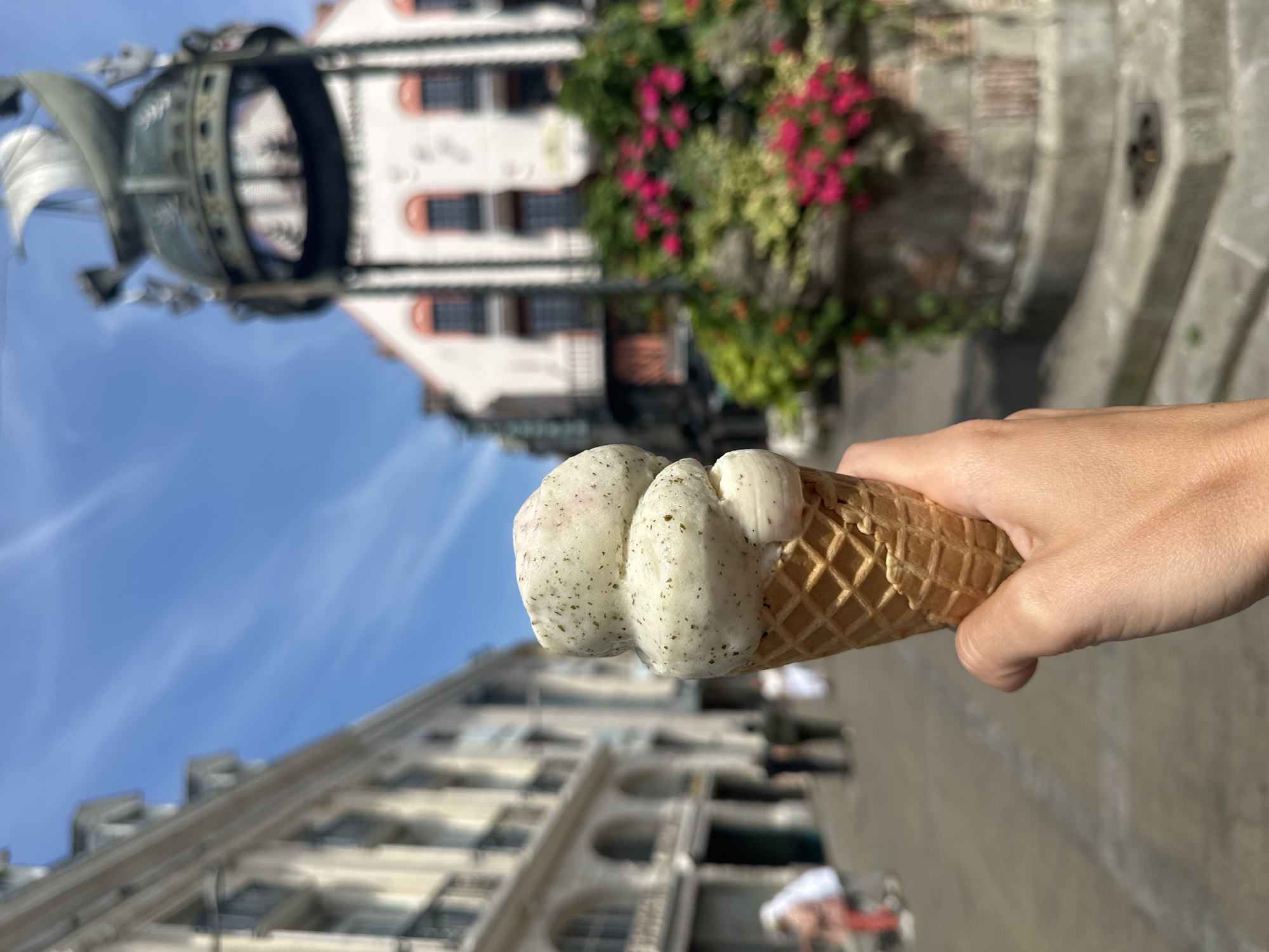 A hand holds a double-scoop ice cream cone in the foreground, with a blurred background of a European city street. The ice cream is a pale green color with dark flecks, likely mint flavored, and has melted slightly. Behind the ice cream, a fountain adorned with pink flowers is visible, and beyond that, a building with arched windows and balconies. The sky is a clear blue with faint wispy clouds. The street appears to be paved, and several blurred figures are walking in the distance. The architecture suggests a historic European city, possibly in France, given the context.