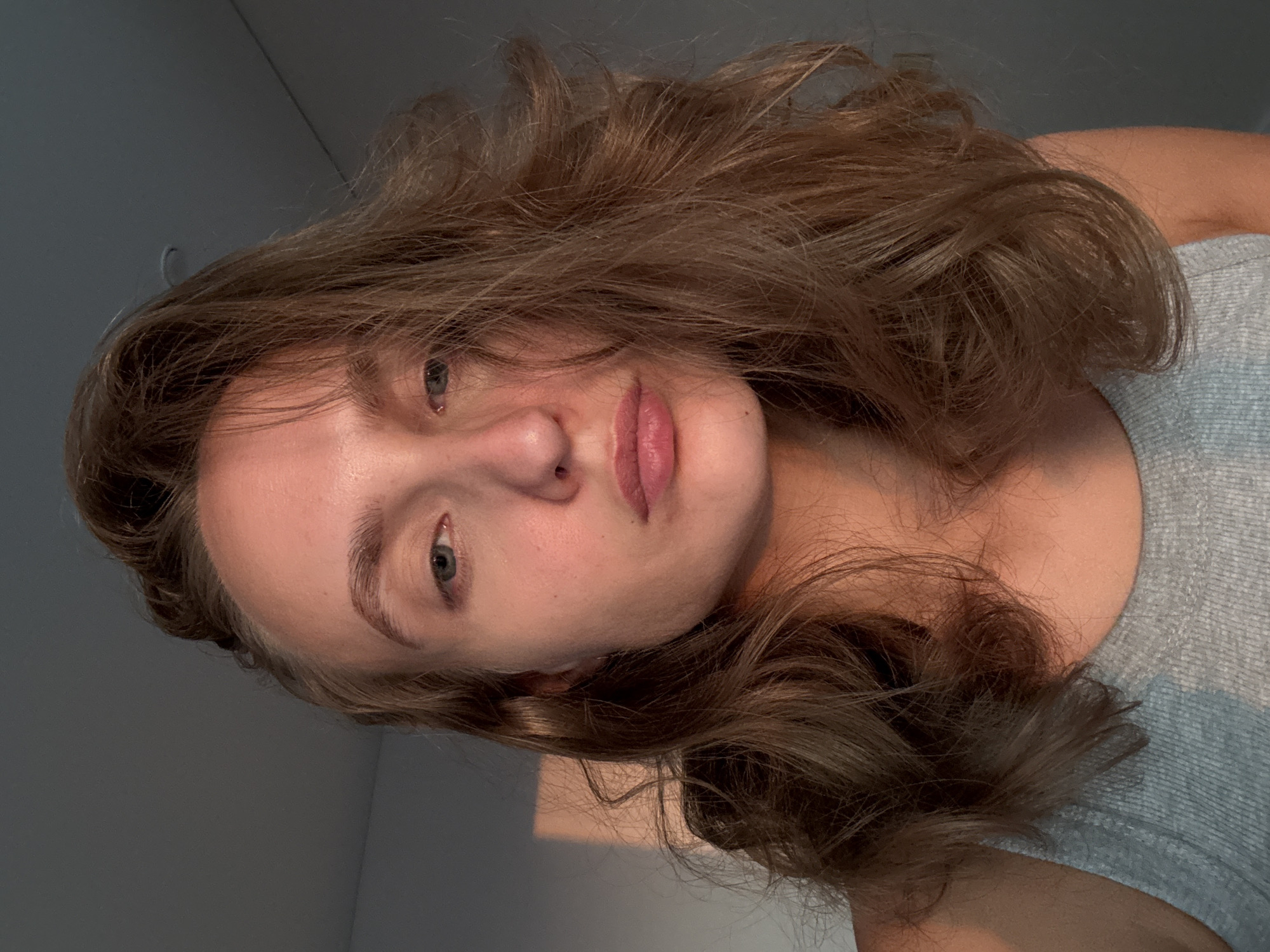 The image is a medium shot portrait of a young Caucasian woman with light brown, wavy hair. She is looking directly at the camera with a neutral expression. Her skin is illuminated by warm, directional light, casting a soft shadow on the left side of her face and a rectangular light pattern on the wall behind her. She is wearing a light gray tank top. The background is a plain, gray wall. The image does not provide any information about the location being in the city of Подшивалово, Russia.