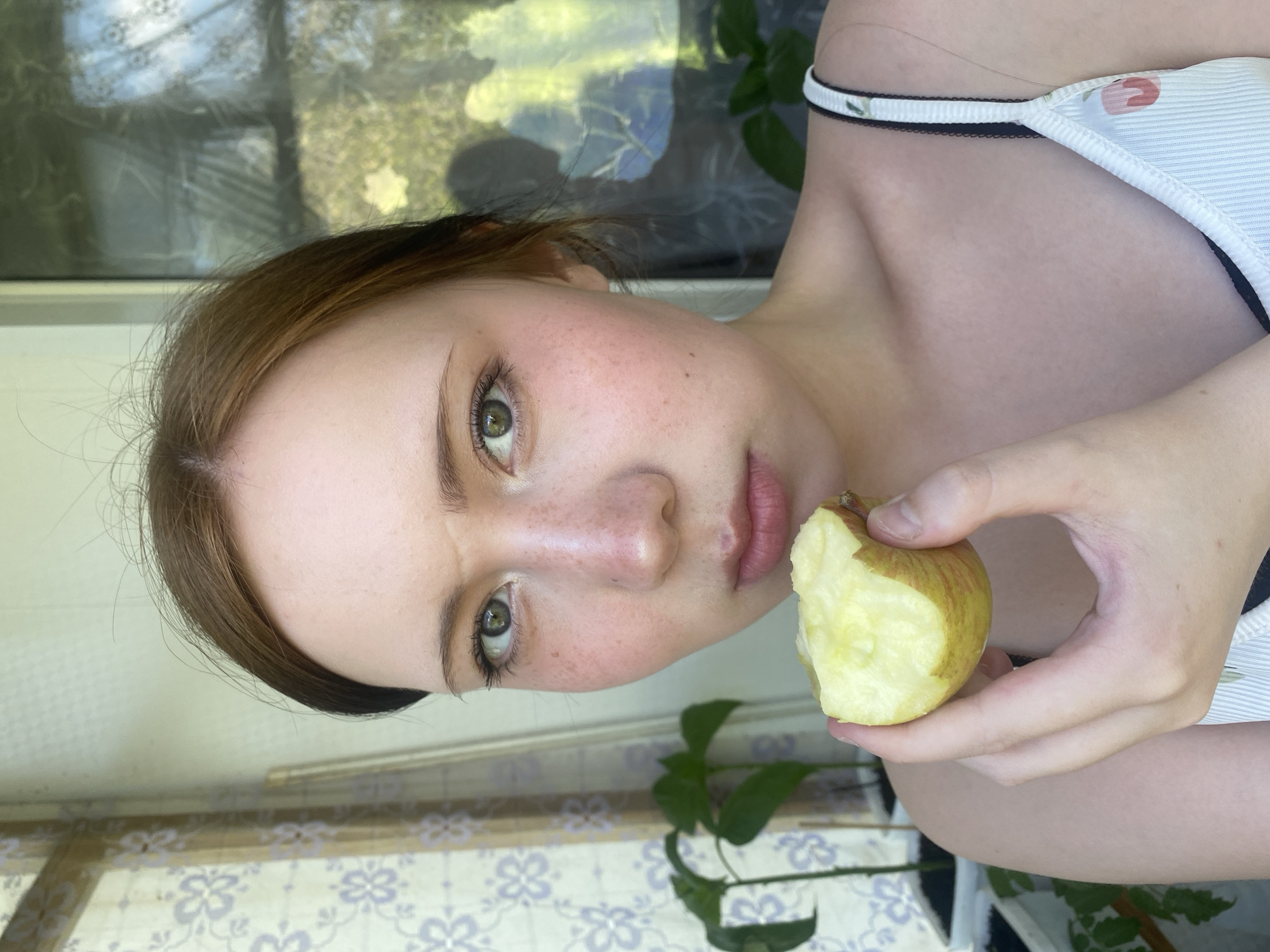 A young woman holds a partially eaten apple in her right hand, looking directly at the camera. She has fair skin, freckles, brown hair, and green eyes. She is wearing a white and black patterned top. The background shows a window with a patterned curtain and indoor plants. The location appears to be indoors.